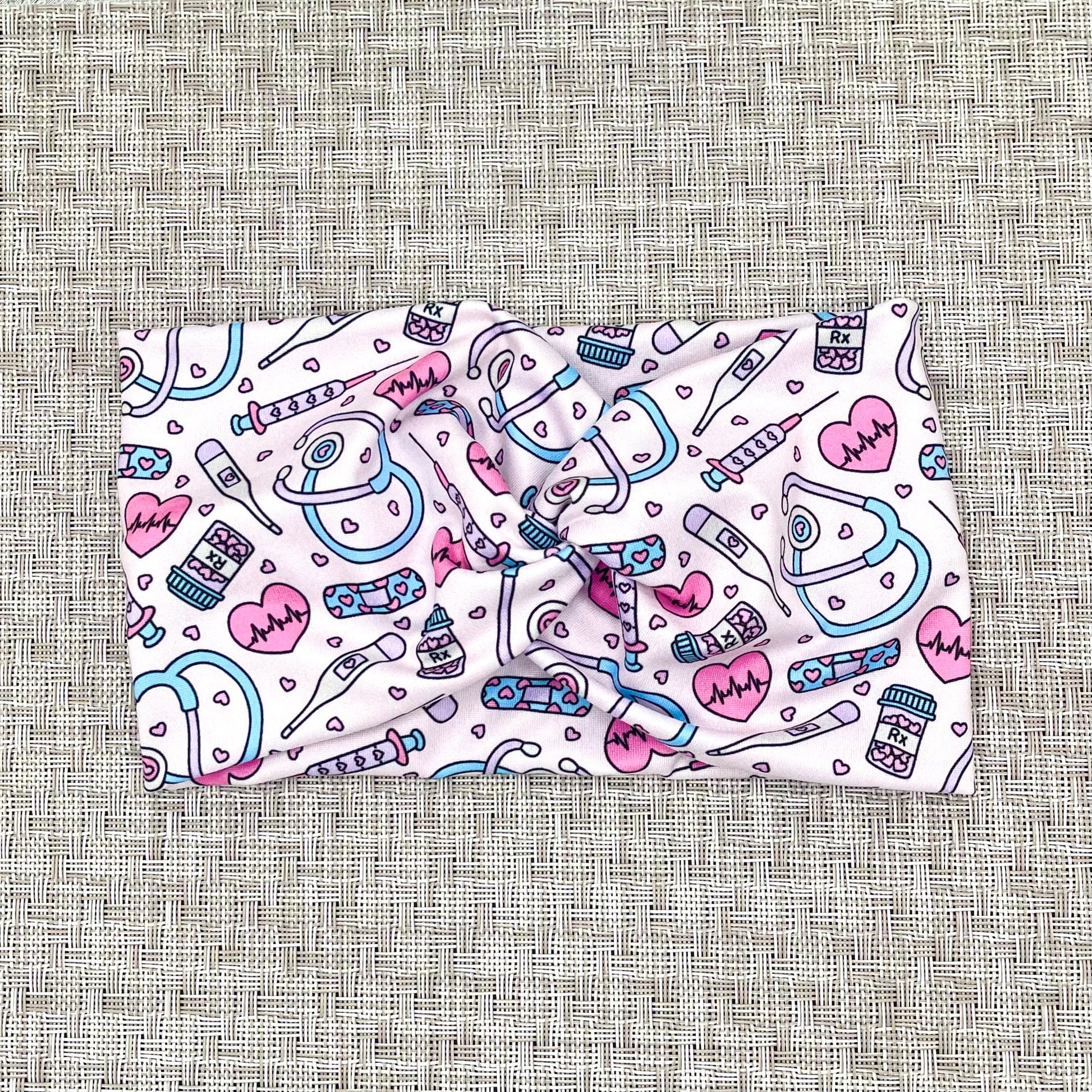 Wide, twisted headband in light pink with pink, purple, and blue medical theme print on a gray background.