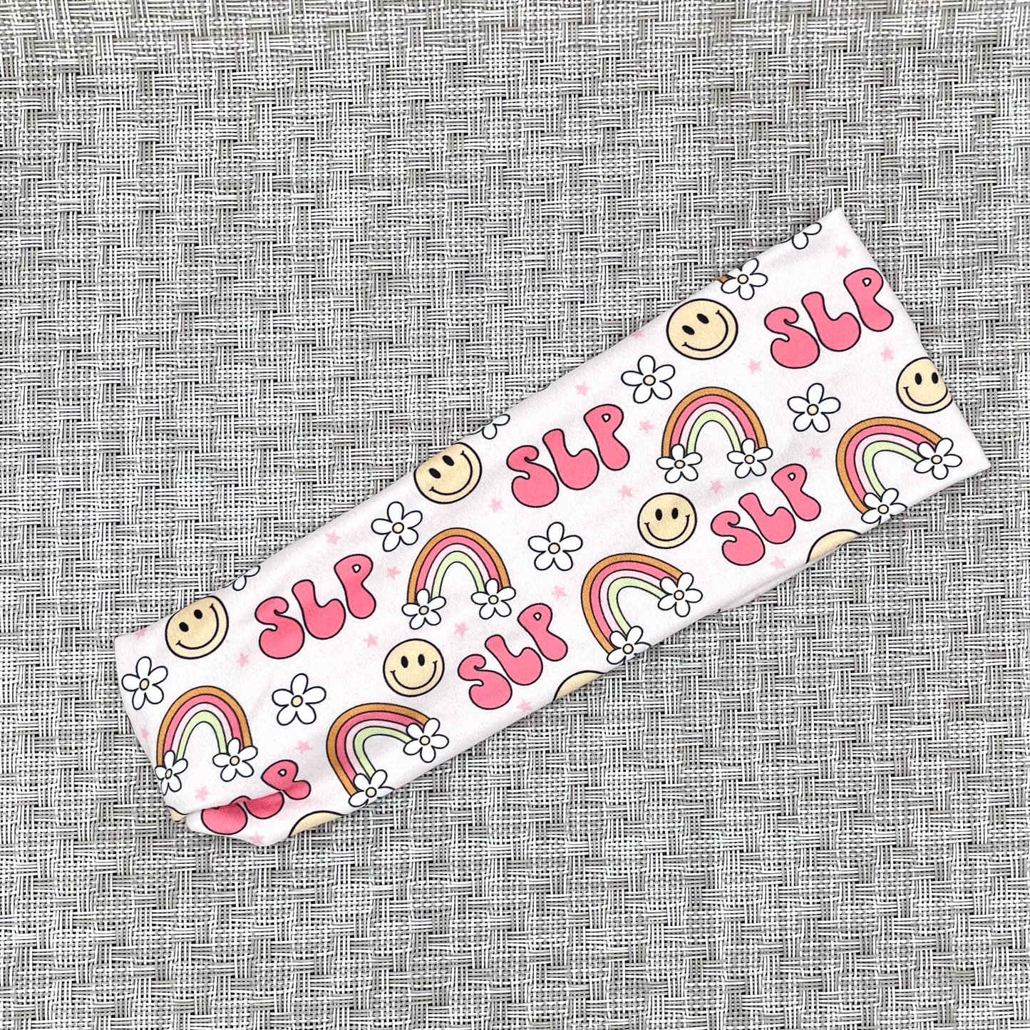 Light pink headband with "SLP" print, rainbows, flowers, and smiley faces.
