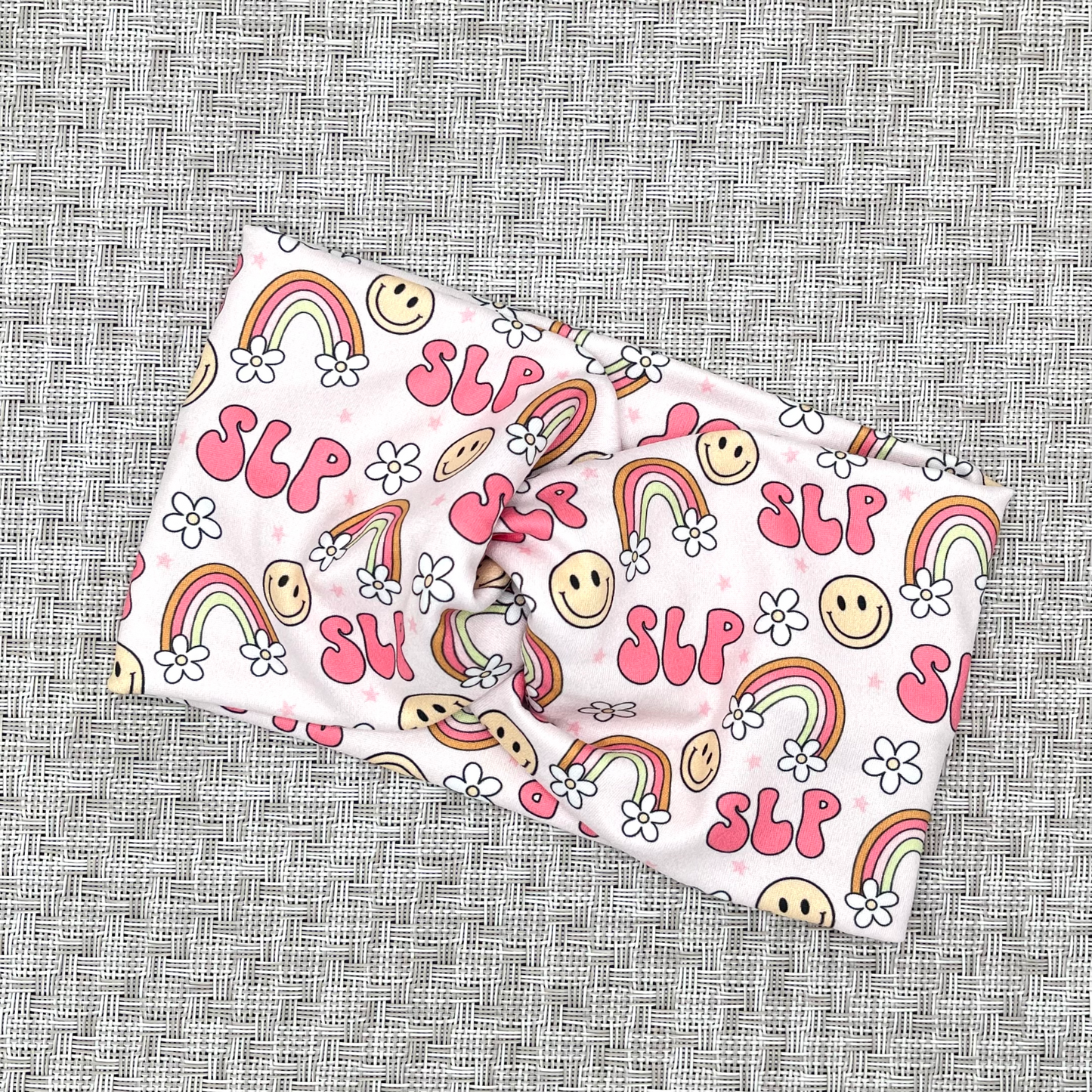 Wide, twisted pink headband stating "slp", with rainbows, smiley faces, and white flowers.