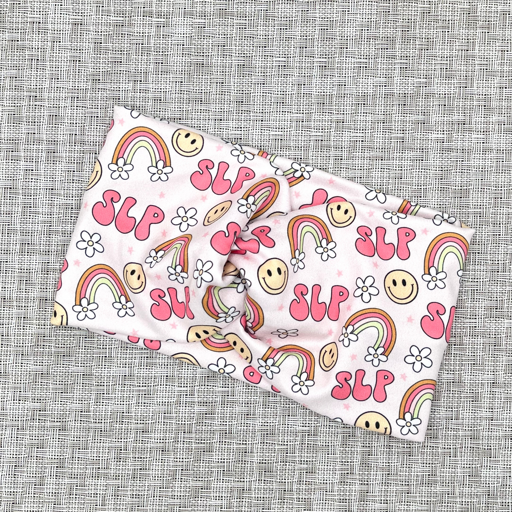 Wide, twisted pink headband stating "slp", with rainbows, smiley faces, and white flowers.