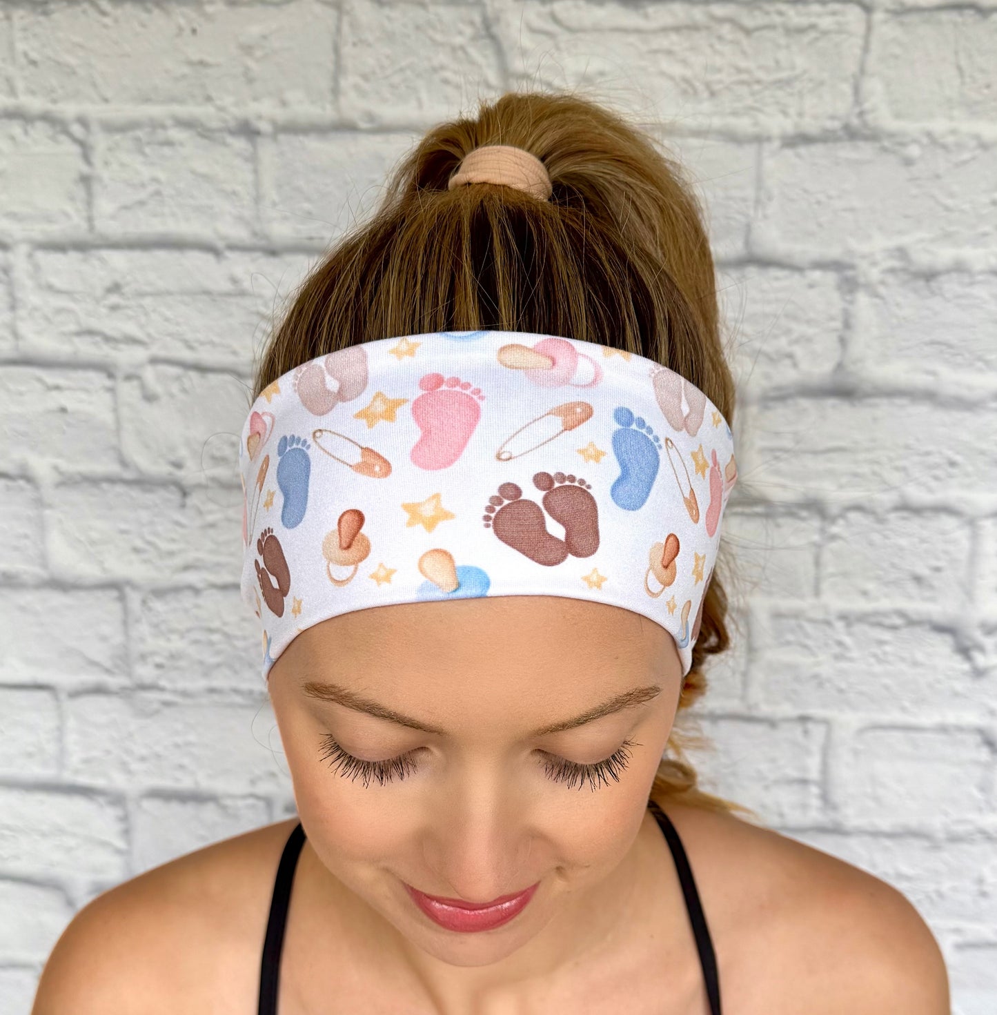 Woman with hair in ponytail weraing flat, stretchy, white headband with baby feet and baby theme print.
