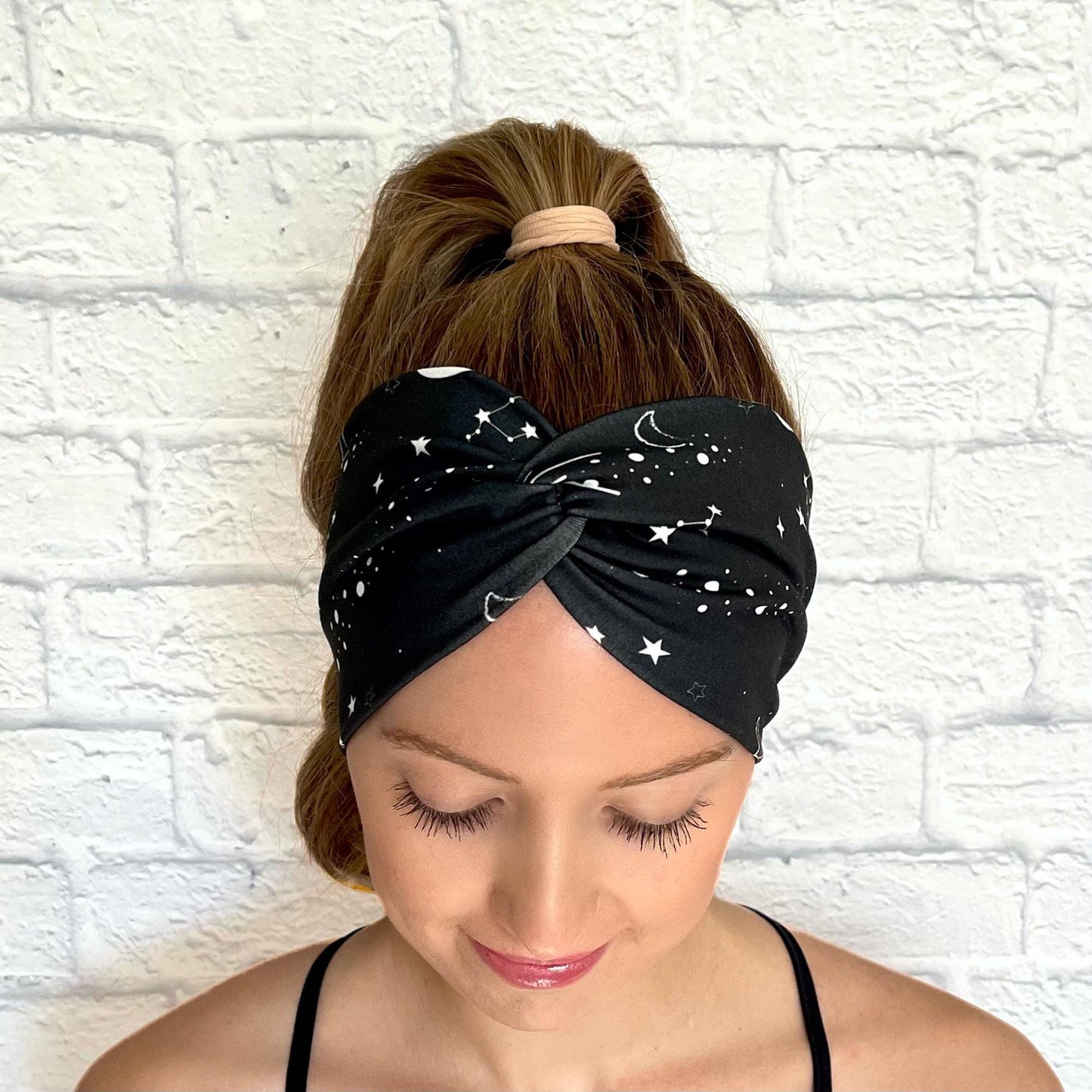 Woman with hair in curled ponytail wearing wide, twisted headband in black with white galaxy theme print.