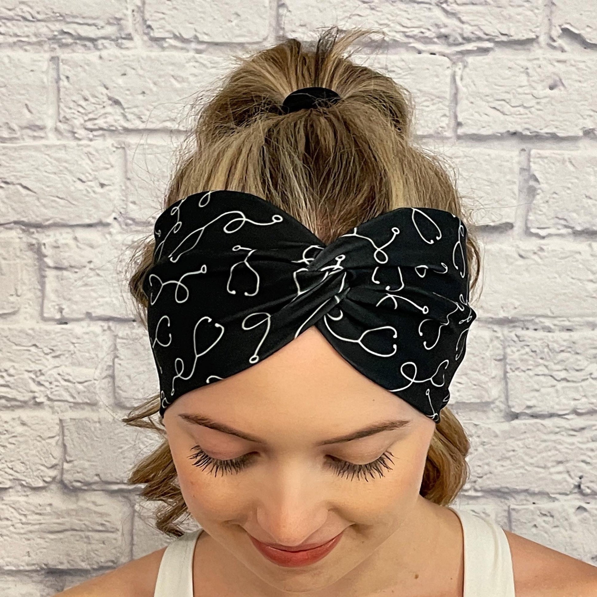 Woman with hair in ponytail wearing black, wide twisted headband with white stethoscope print.
