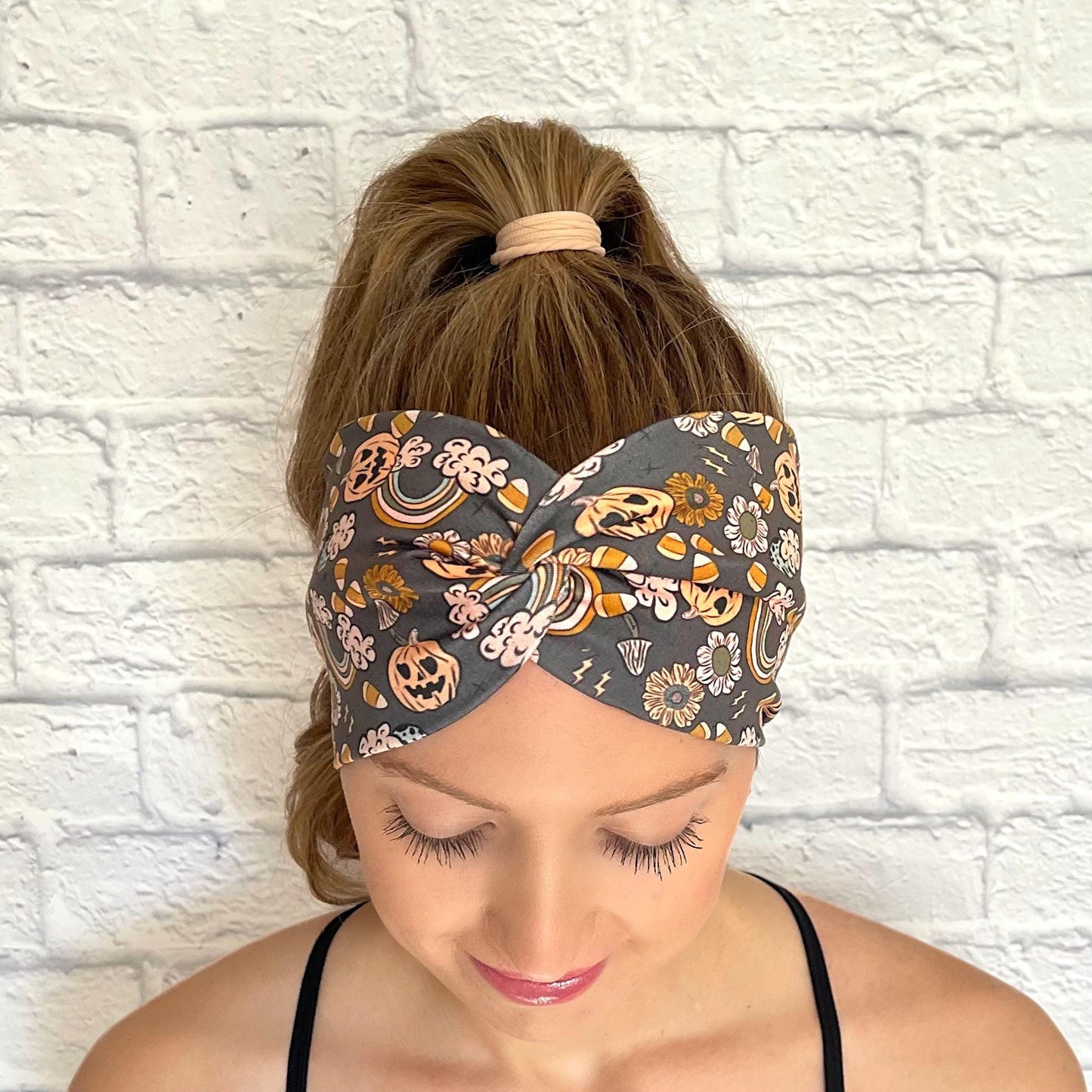 Woman with hair in curled ponytail wearing wide, twisted headband with orange Halloween theme print- pumpkins, rainbows, candy corn, and flowers.