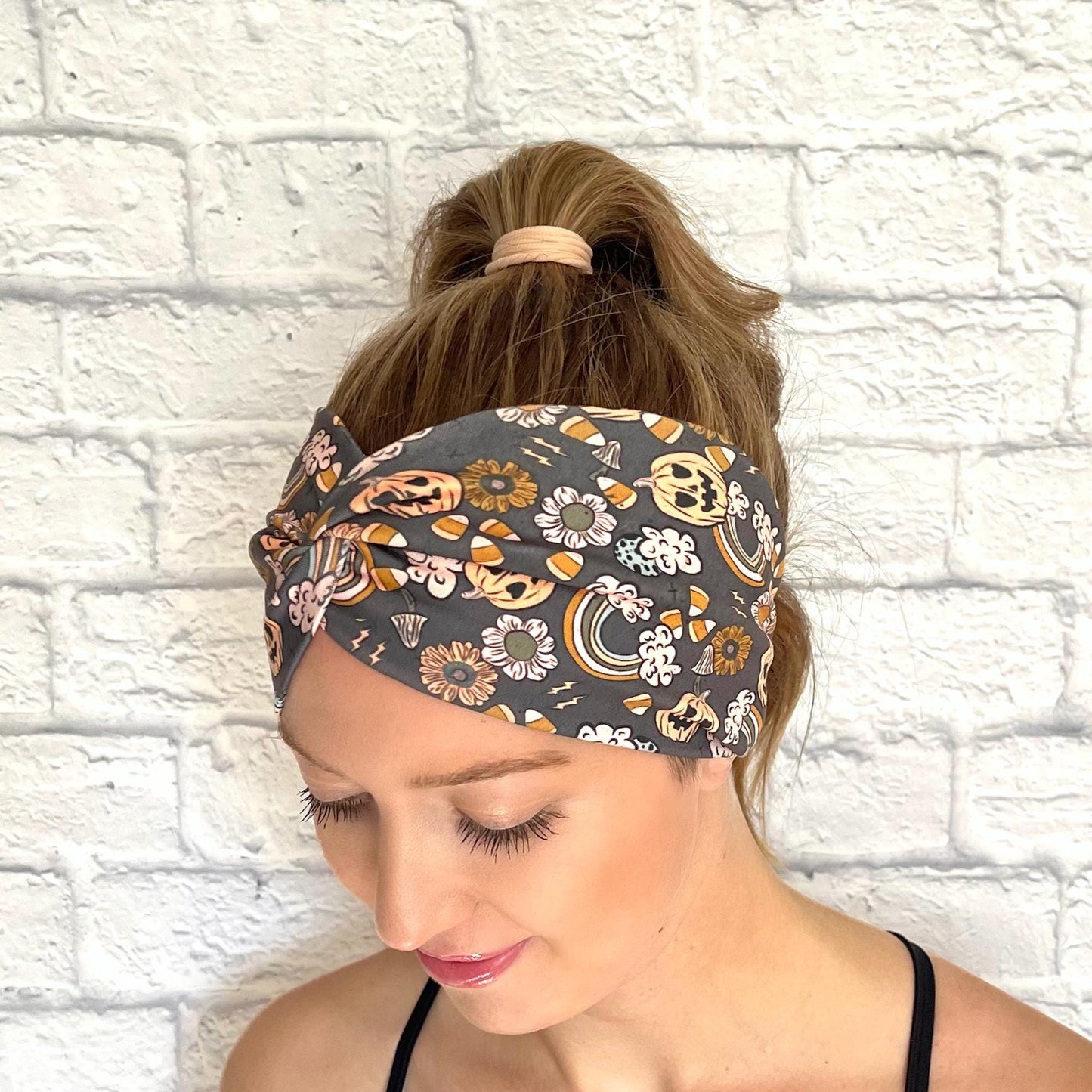 Woman with hair in curled ponytail wearing wide, twisted headband with orange Halloween theme print- pumpkins, rainbows, candy corn, and flowers.