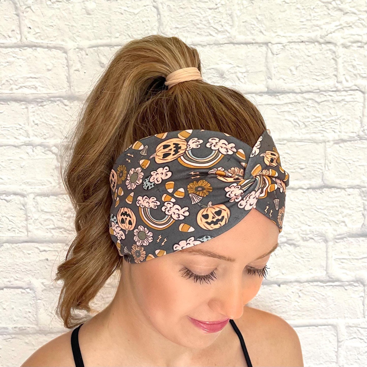 Woman with hair in curled ponytail wearing wide, twisted headband with orange Halloween theme print- pumpkins, rainbows, candy corn, and flowers.