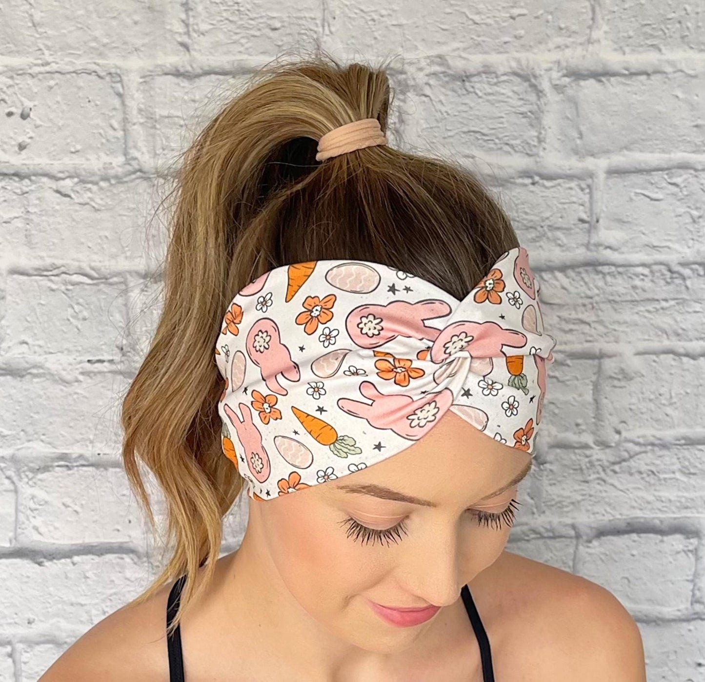 Woman with hair in curled ponytail wearing wide, twisted headband in white with orange and peach Easter theme print.
