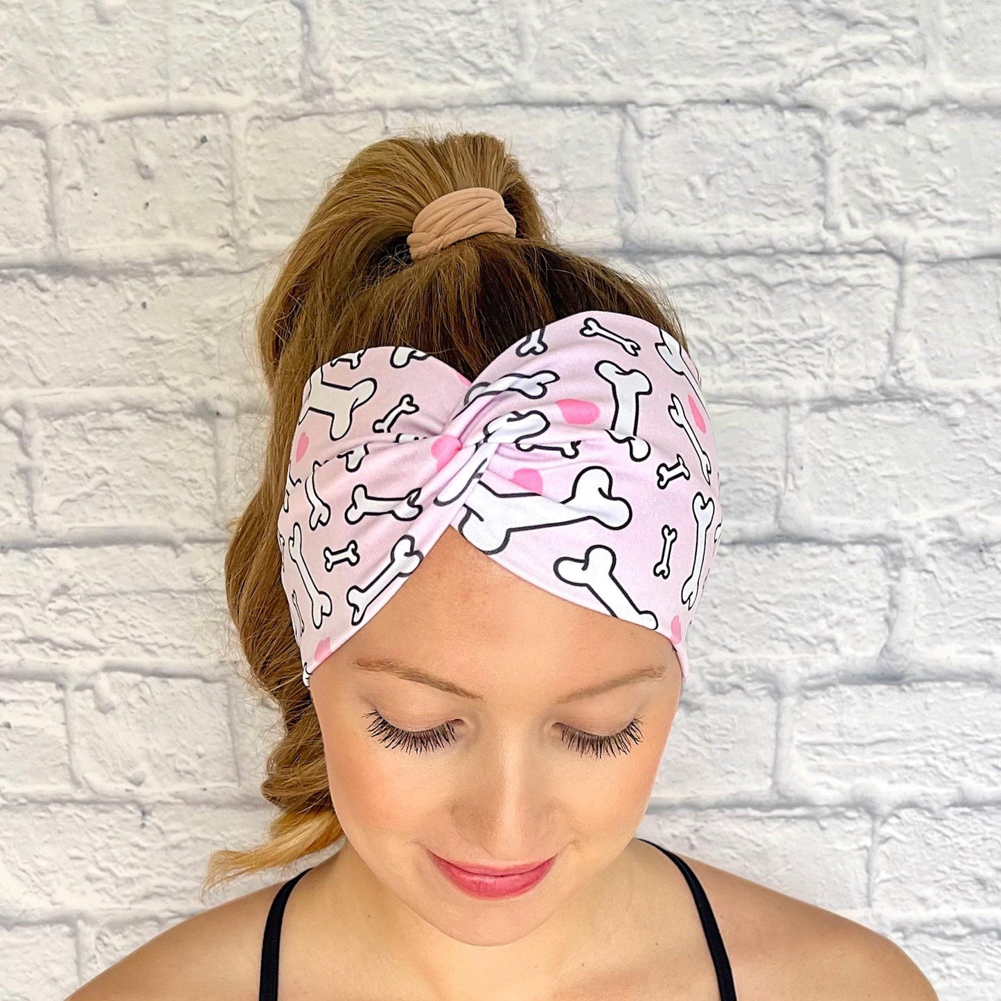 Woman with hair in curled ponytail wearing wide, twisted headband in light pink with dog bone and heart print.