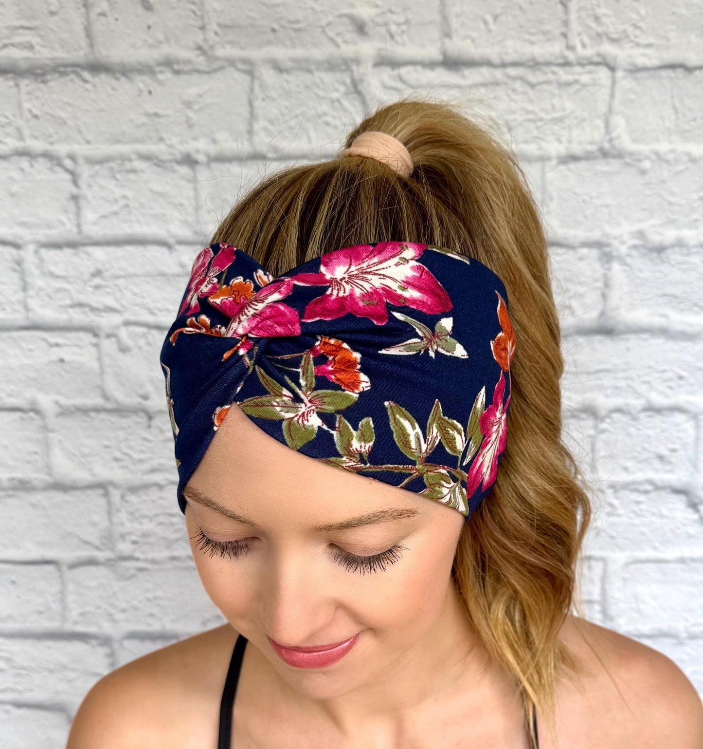 Woman with hair in ponytail wearing wide, twisted, navy color headband with pink, orange, and green floral print.