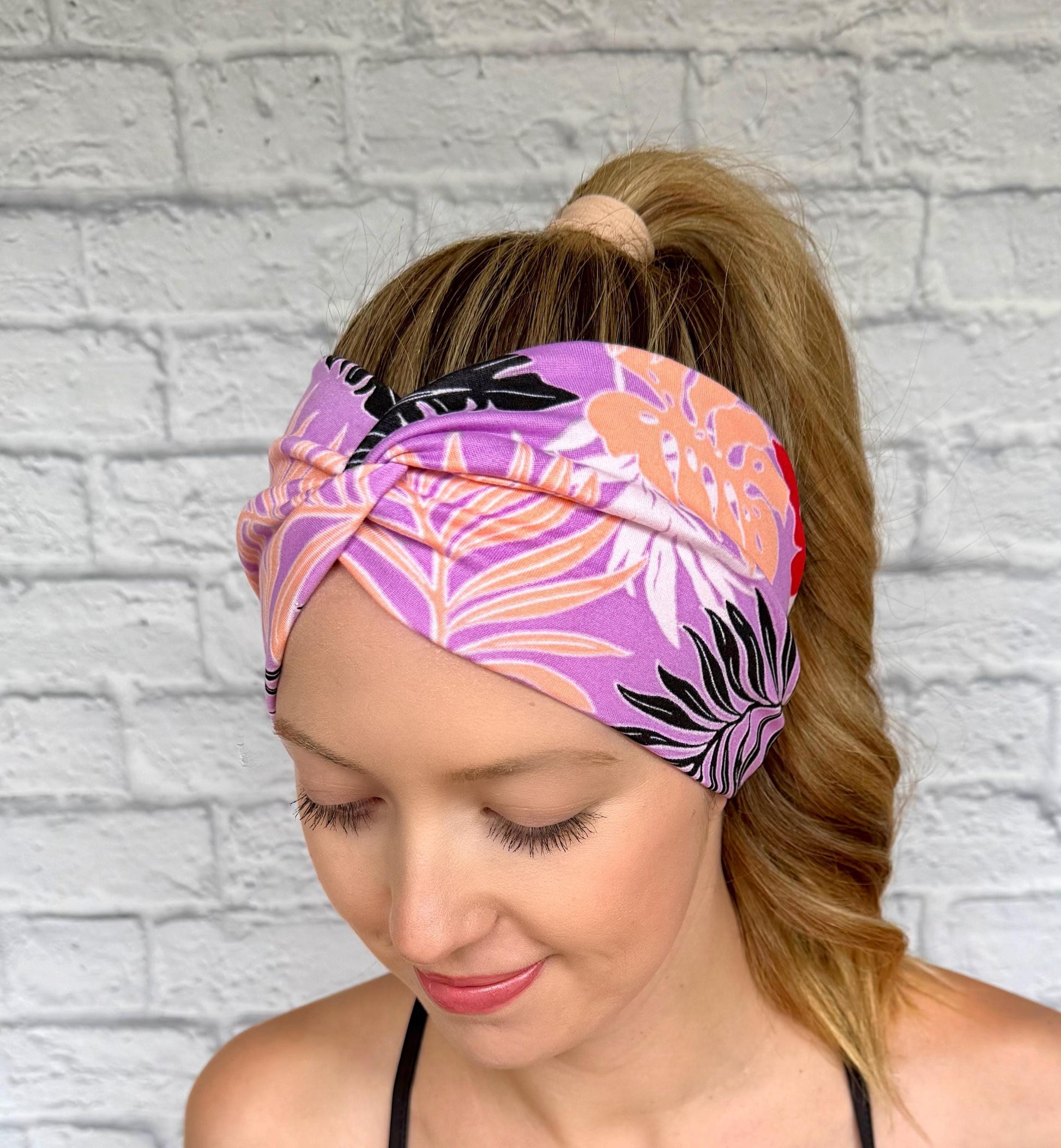 Woman with hair in ponytail wearing wide, twisted headband in lavender color with black, peach, white, and red floral print.