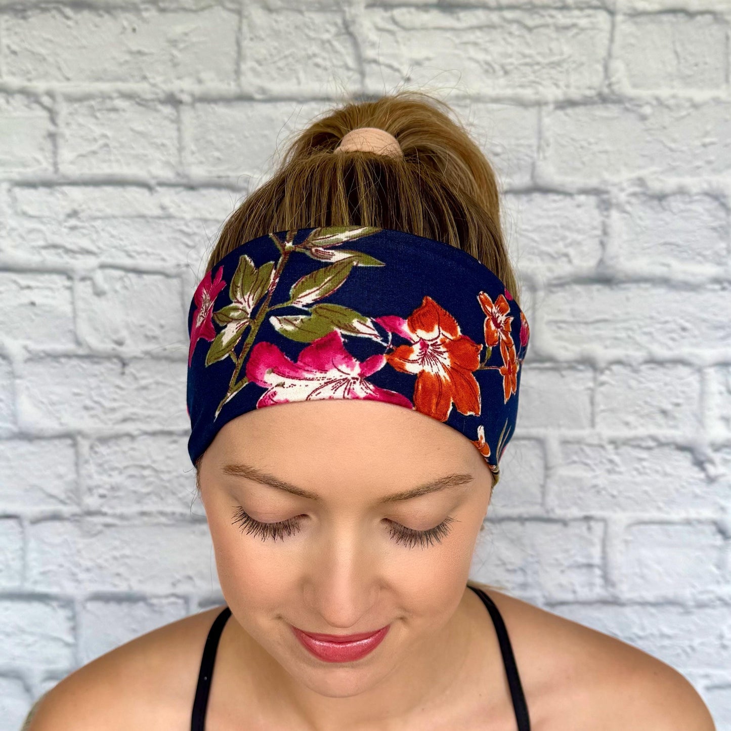 Woman with hair in ponytail wearing flat, stretchy, navy color headband with floral print, primarily pink florals.