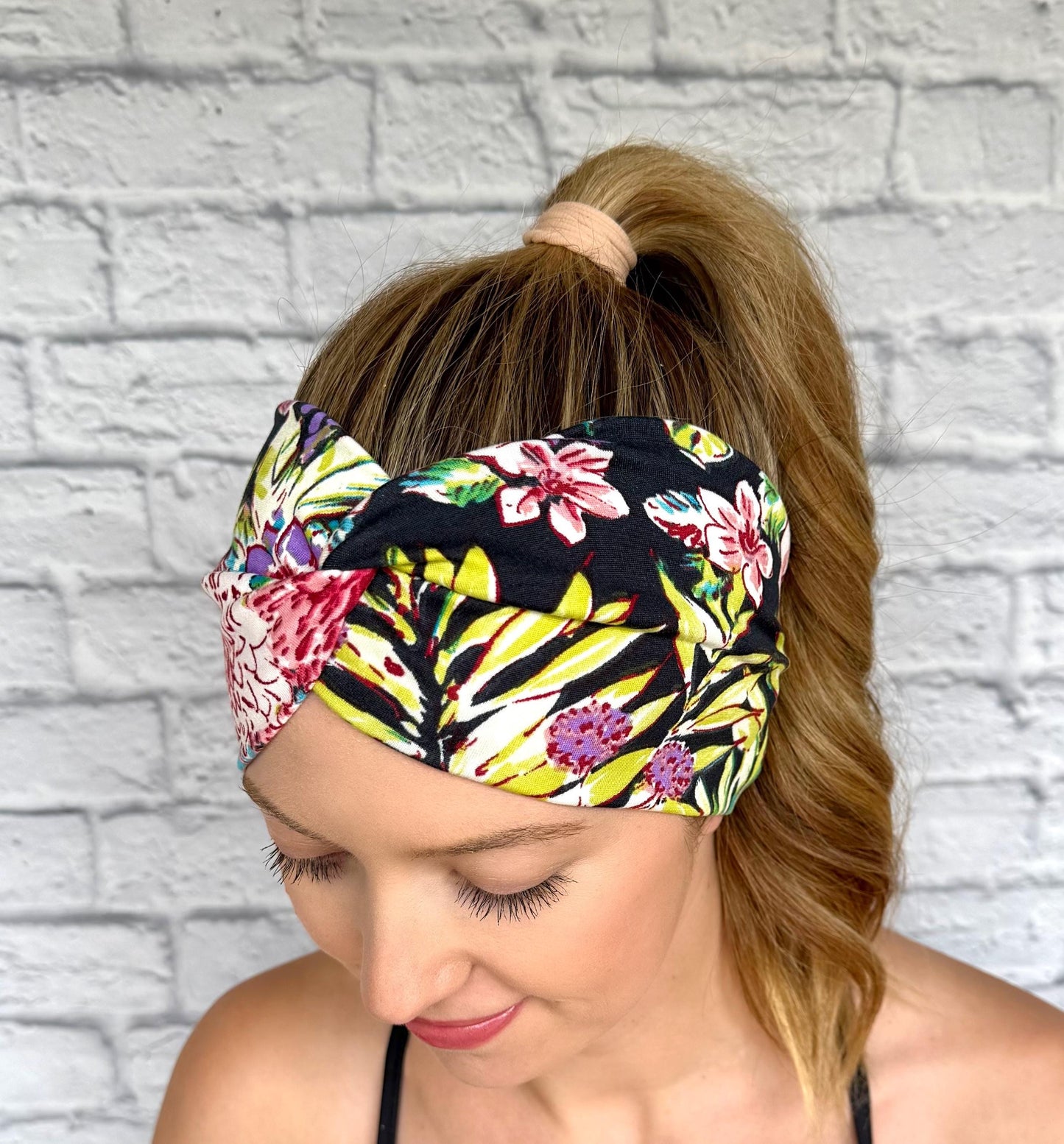 Woman with hair in ponytail wearing wide, twisted headband in black with lime green, pink, white, and purple floral print.