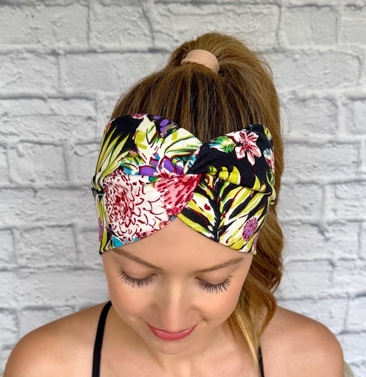 Woman with hair in ponytail wearing wide, twisted headband in black with lime green, pink, white, and purple floral print.