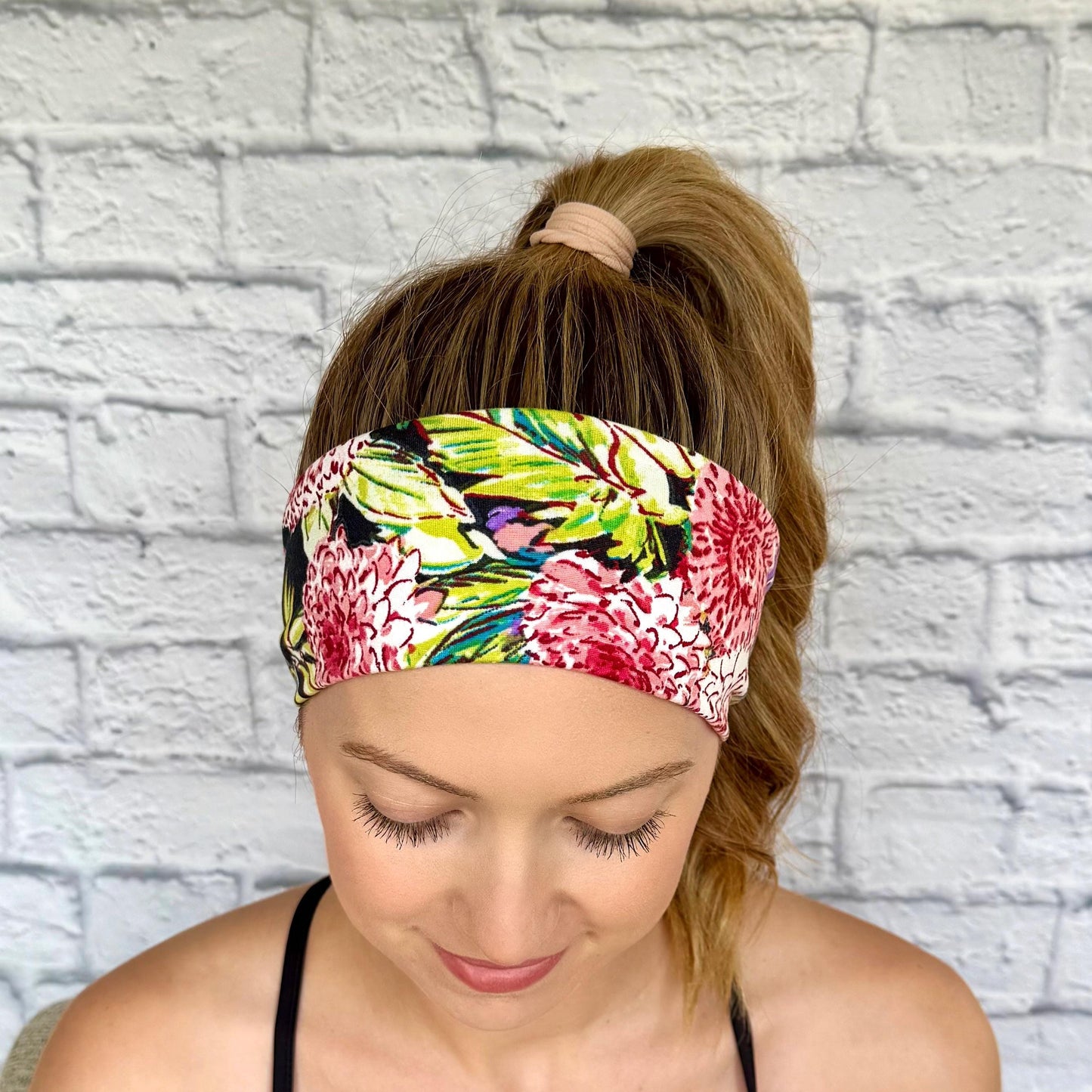 Woman with hair in ponytail wearing flat, stretchy headband in black with lime green, pink, and white floral print.