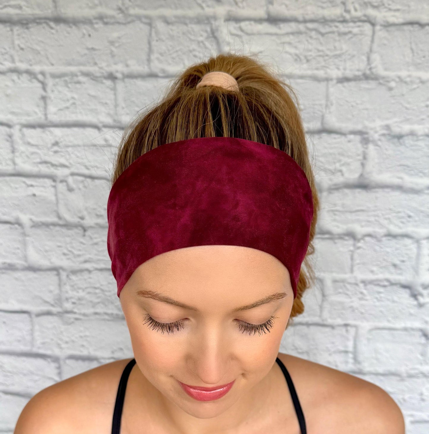 Woman with hair in ponytail wearing flat, stretchy headband in maroon color tie dye print.