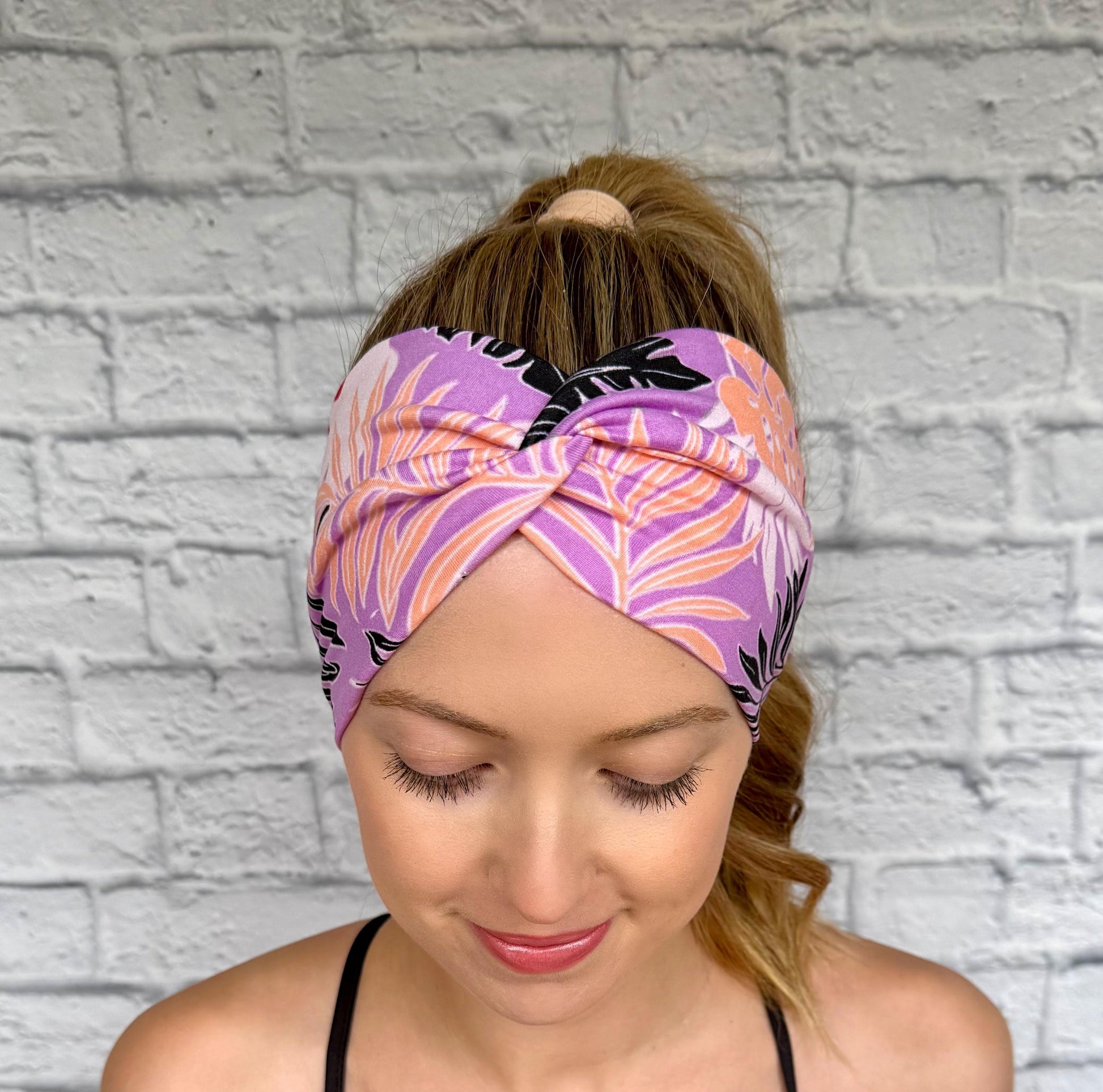 Woman with hair in ponytail wearing wide, twisted headband in lavender color with black, peach, white, and red floral print.