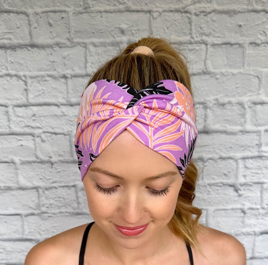 Woman with hair in ponytail wearing wide, twisted headband in lavender color with black, peach, white, and red floral print.