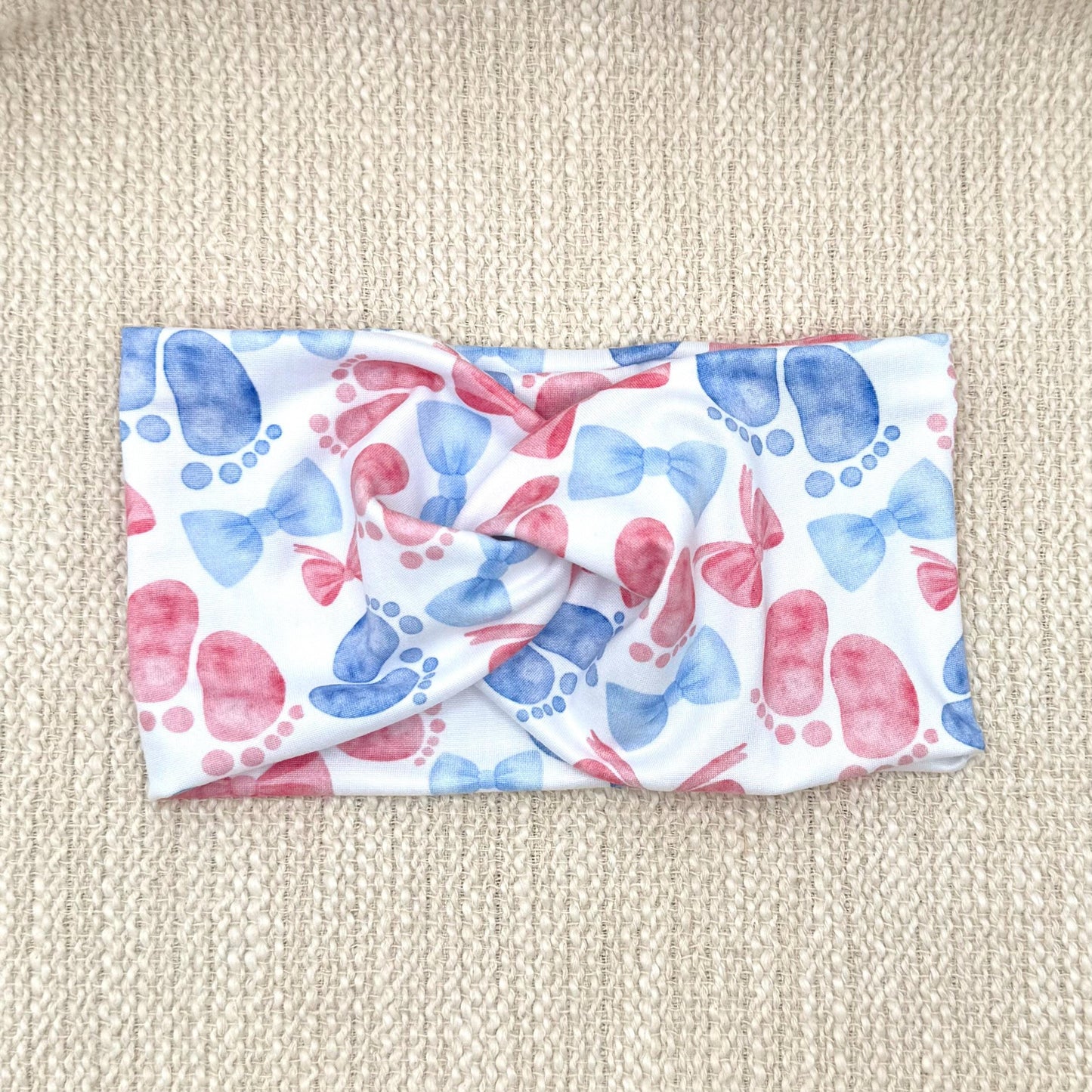 Wide, twisted, white headband with pink and blue baby feet and bow print.