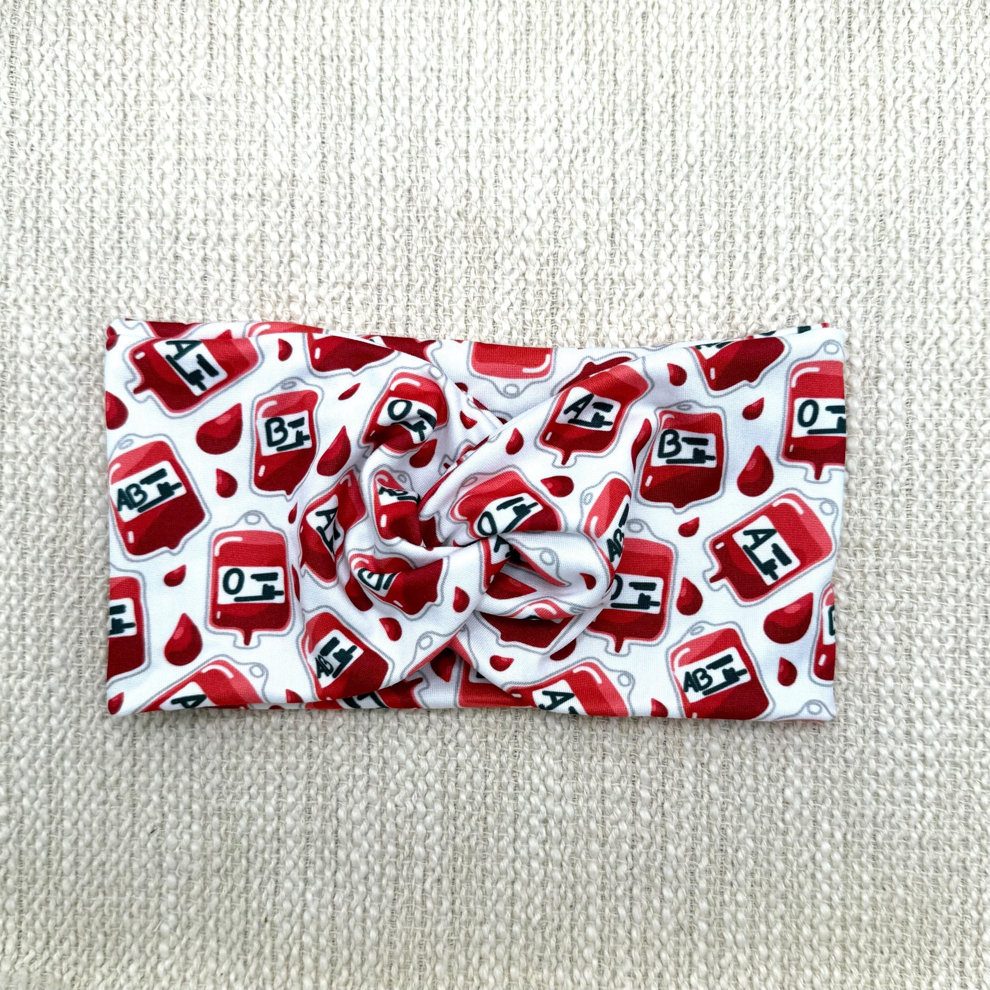Wide, twisted headband in white with blood/ phlebotomy theme print displayed on a cream color background.