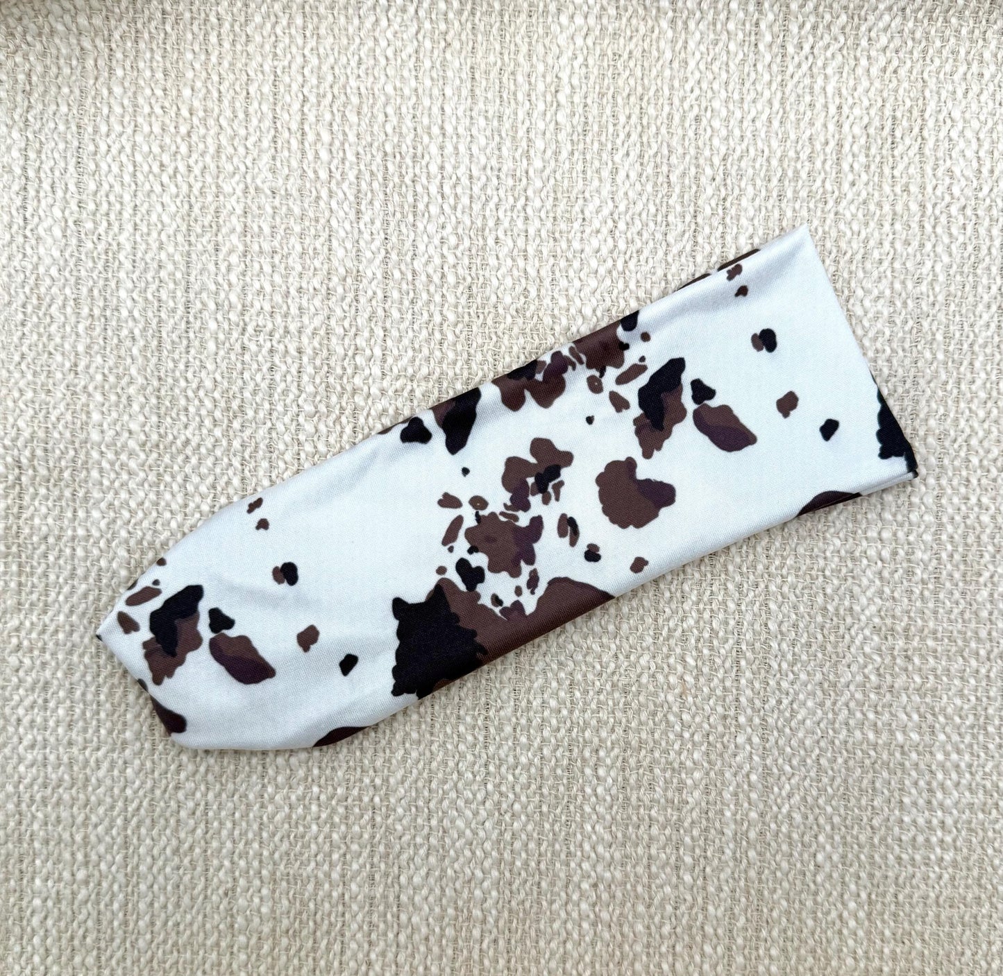 Flat, stretchy headband in white with cow print displayed on a cream color background.