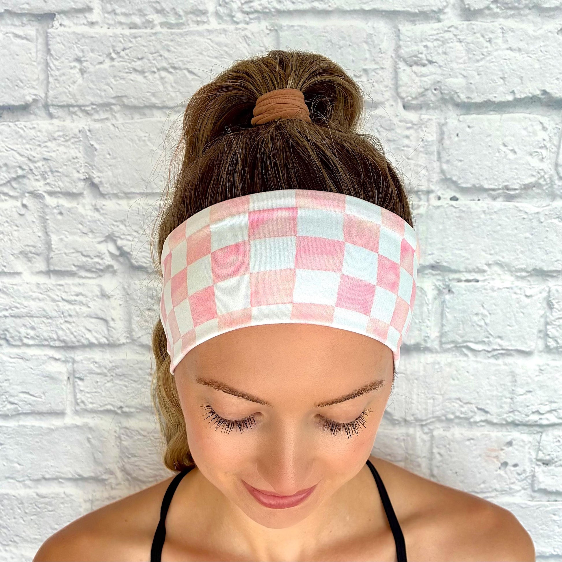 Woman with hair in ponytail wearing flat, stretchy headband in pink and white checker print.