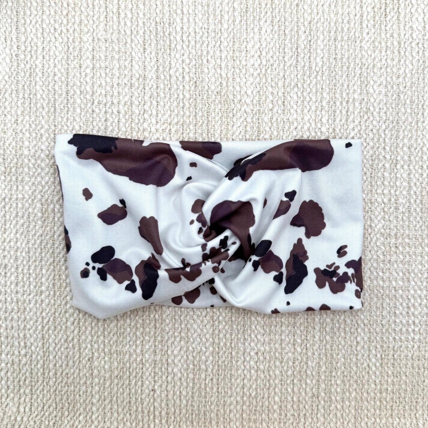 Wide, twisted headband with cow print displayed on a cream color background.
