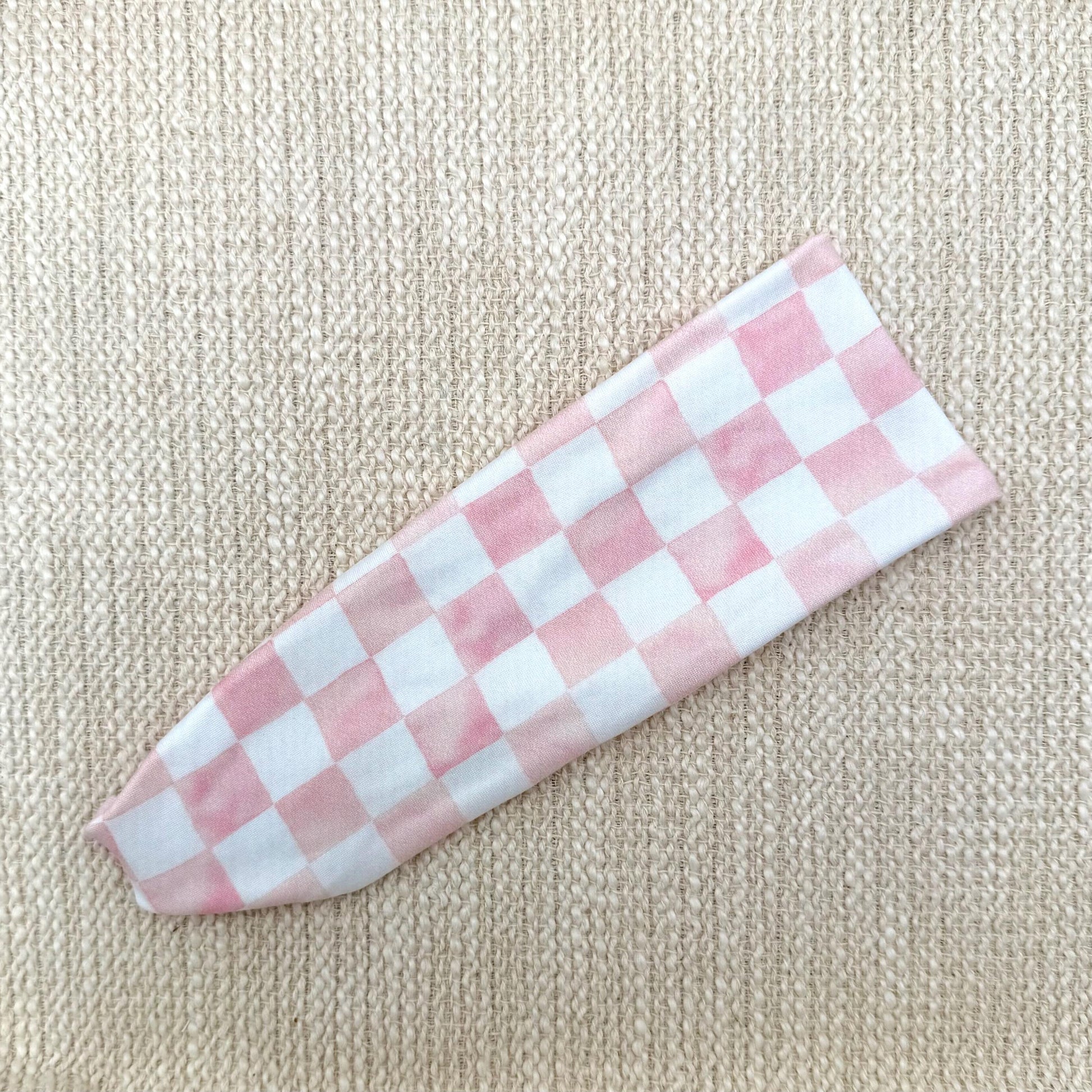Flat, stretchy headband in pink and white checker print displayed on a cream color background.