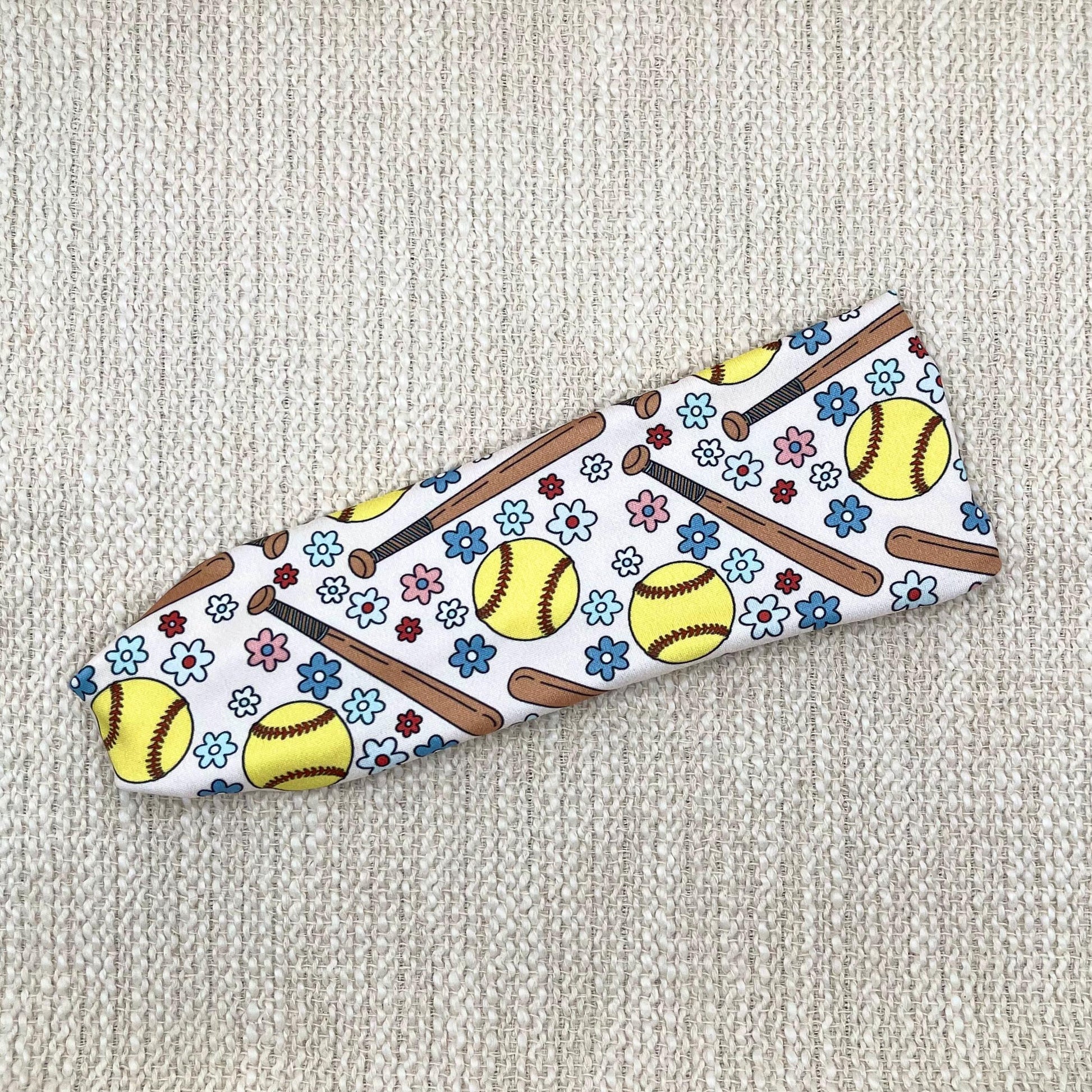 Flat, stretchy headband in cream color with softball and flower theme print.