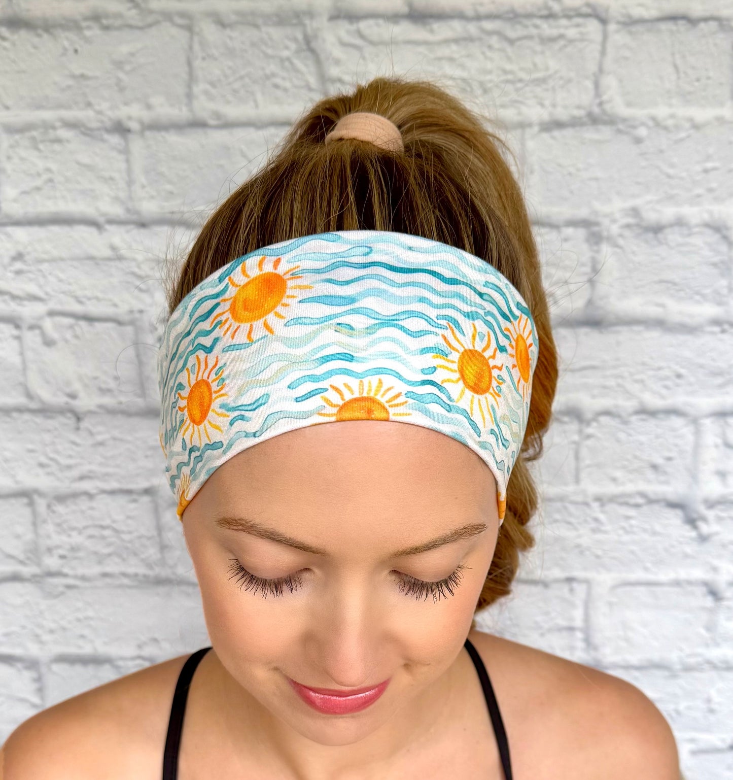 Woman with hair in ponytail wearing flat, stretchy headband with blue waves and sun print.