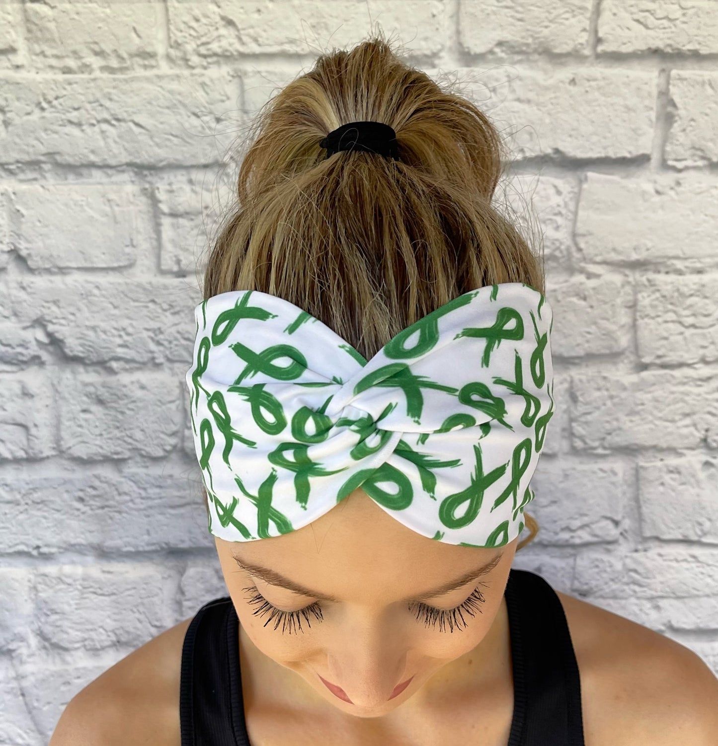 Woman with hair in ponytail wearing wide, twisted, white headband with green ribbon print.