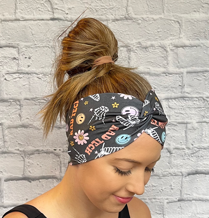 Woman with hair in bun wearing a dark gray, wide, twisted headband with skeletons, flowers, smiley faces, and "rad tech" print.