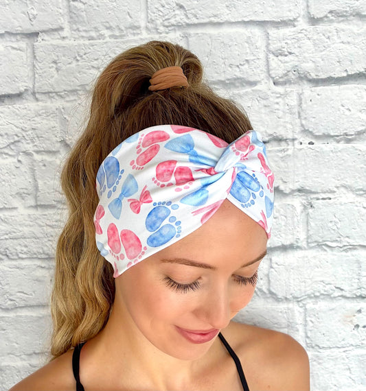 Woman with hair in ponytail wearing wide, twisted, white headband with pink and blue baby feet and bow print.