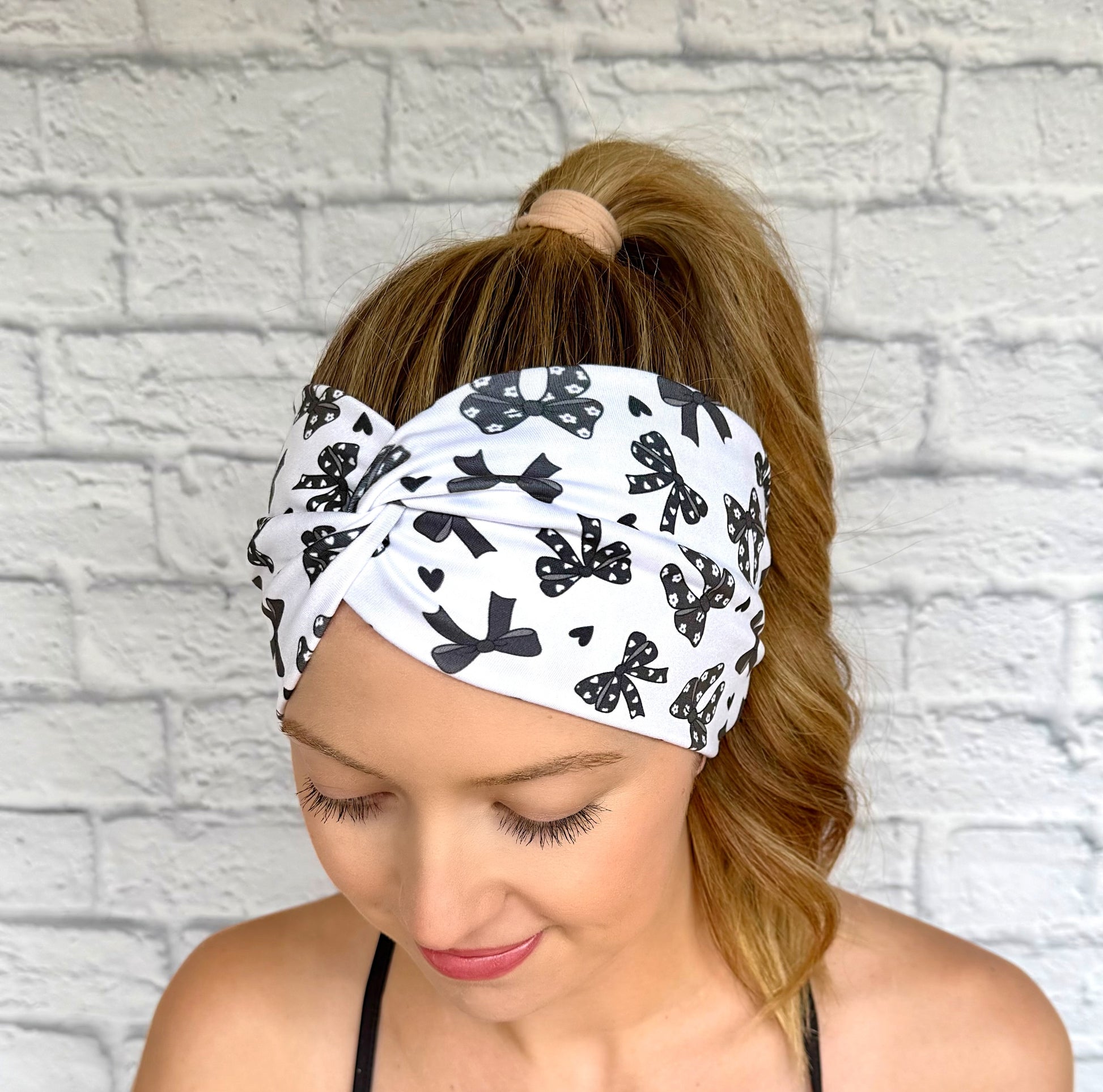 Woman with hair in curled ponytail wearing wide, twisted, white headband with black bow print.