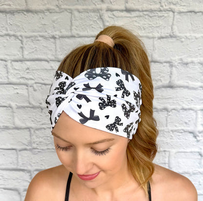 Woman with hair in curled ponytail wearing wide, twisted, white headband with black bow print.