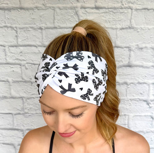 Woman with hair in curled ponytail wearing wide, twisted, white headband with black bow print.
