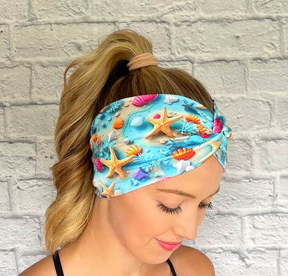 Woman with hair in ponytail wearing light blue, wide twist headband with ocean theme print- water, sand, starfish, and shells