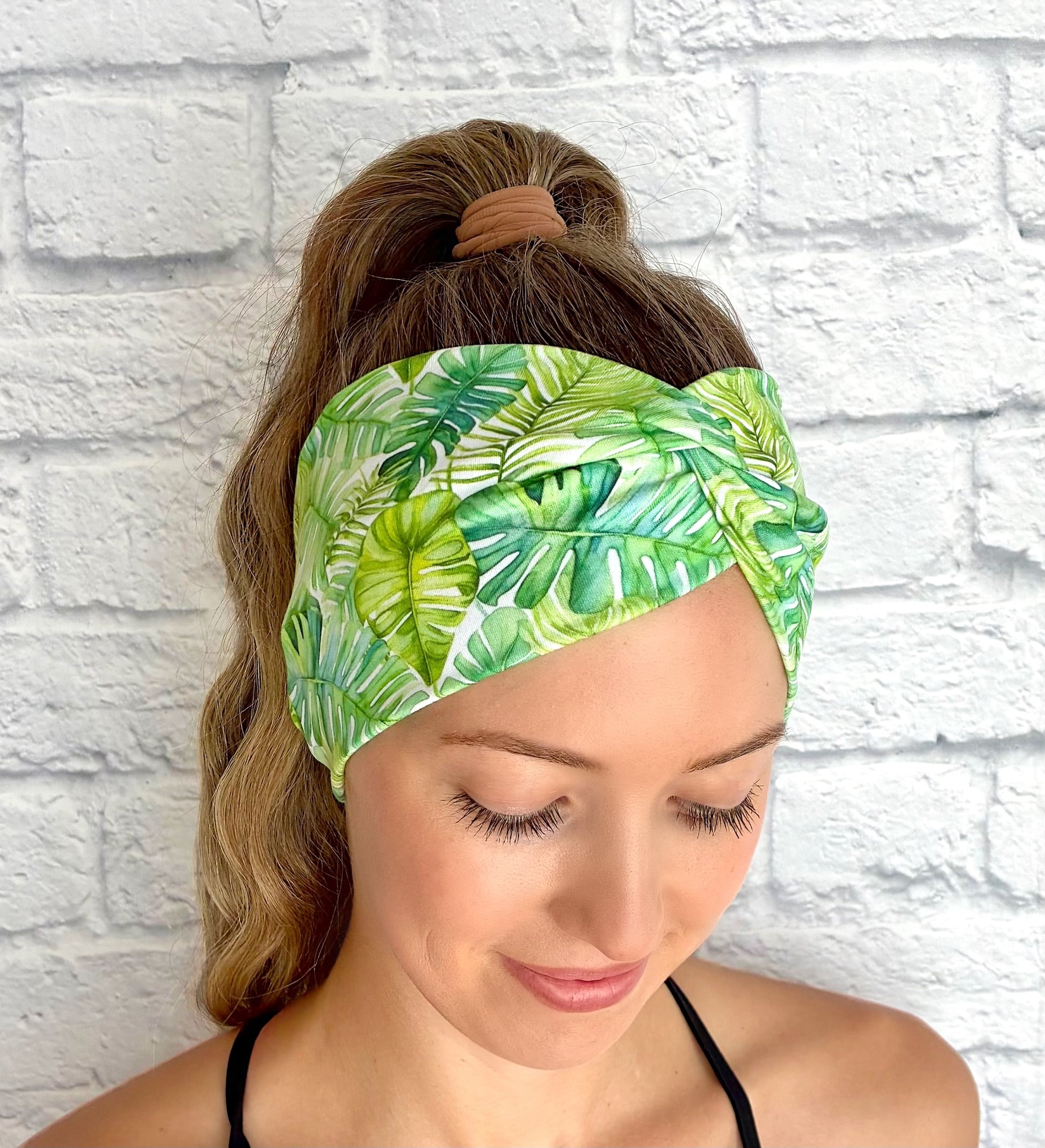 Woman wearing a green palm leaf-patterned headband against a white brick wall.
