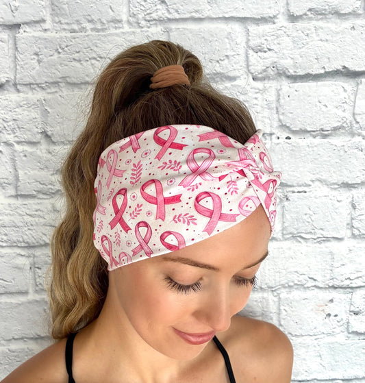 Woman with hair in ponytail wearing wide, twisted, white/cream headband with pink ribbon and pink floral print against a white brick wall. 