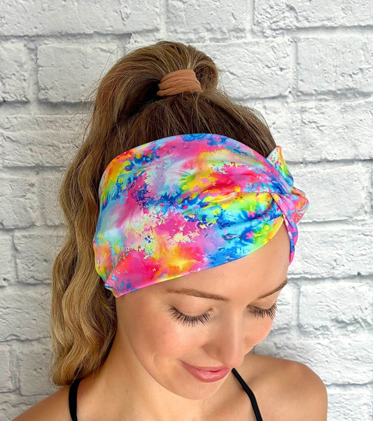 Woman wearing wide, twisted headband in rainbow color paint-splatter looking print.