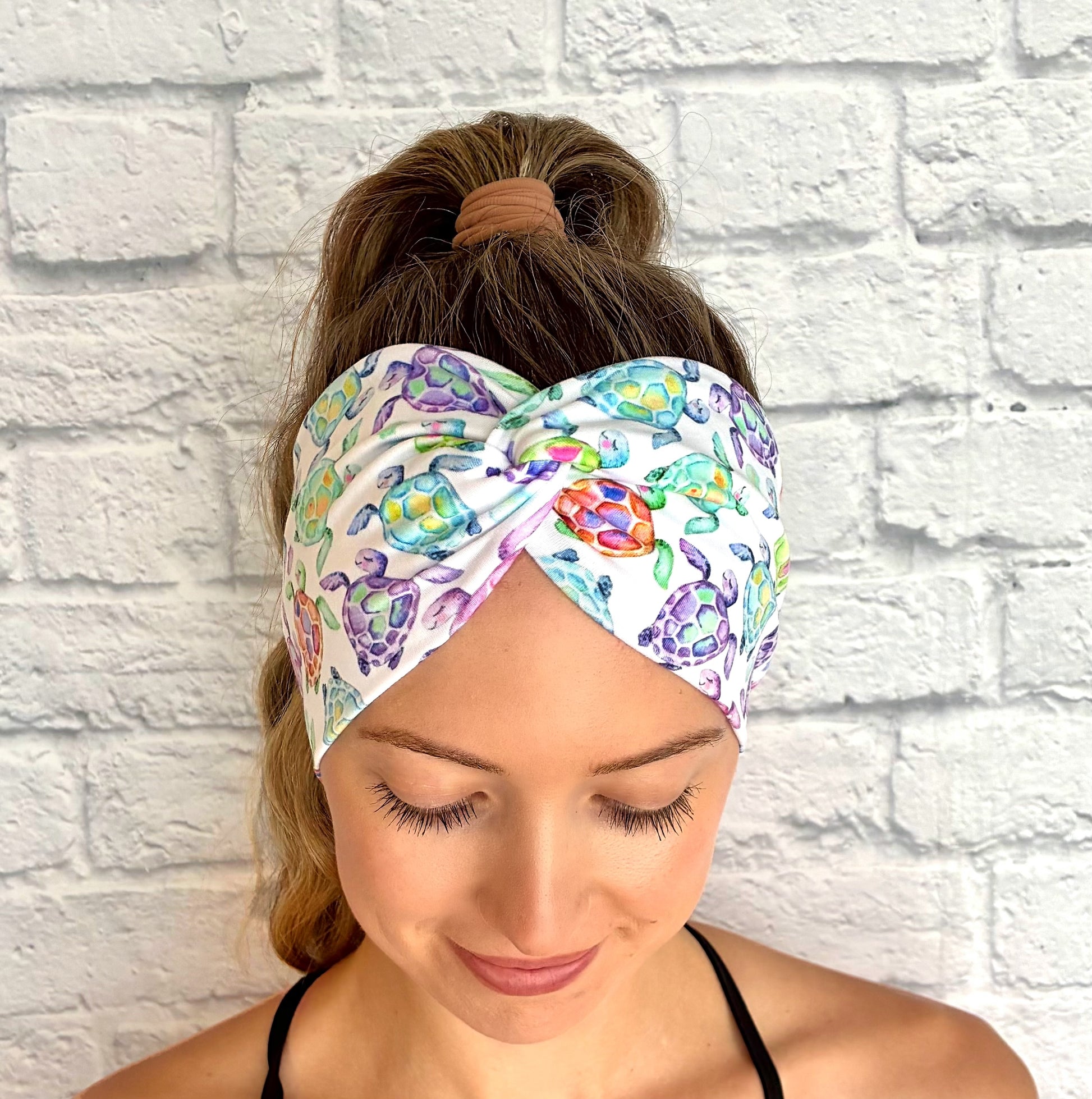 Woman with hair in ponytail wearing wide, twisted, white headband with sea turtle print, displayed on white brick background.