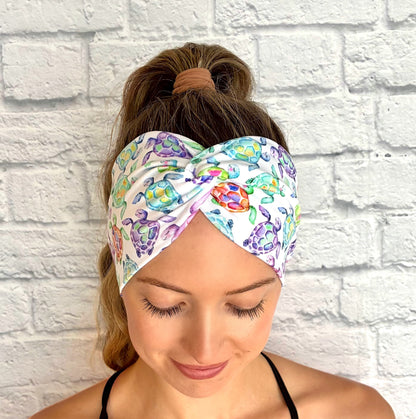 Woman with hair in ponytail wearing wide, twisted, white headband with sea turtle print, displayed on white brick background.