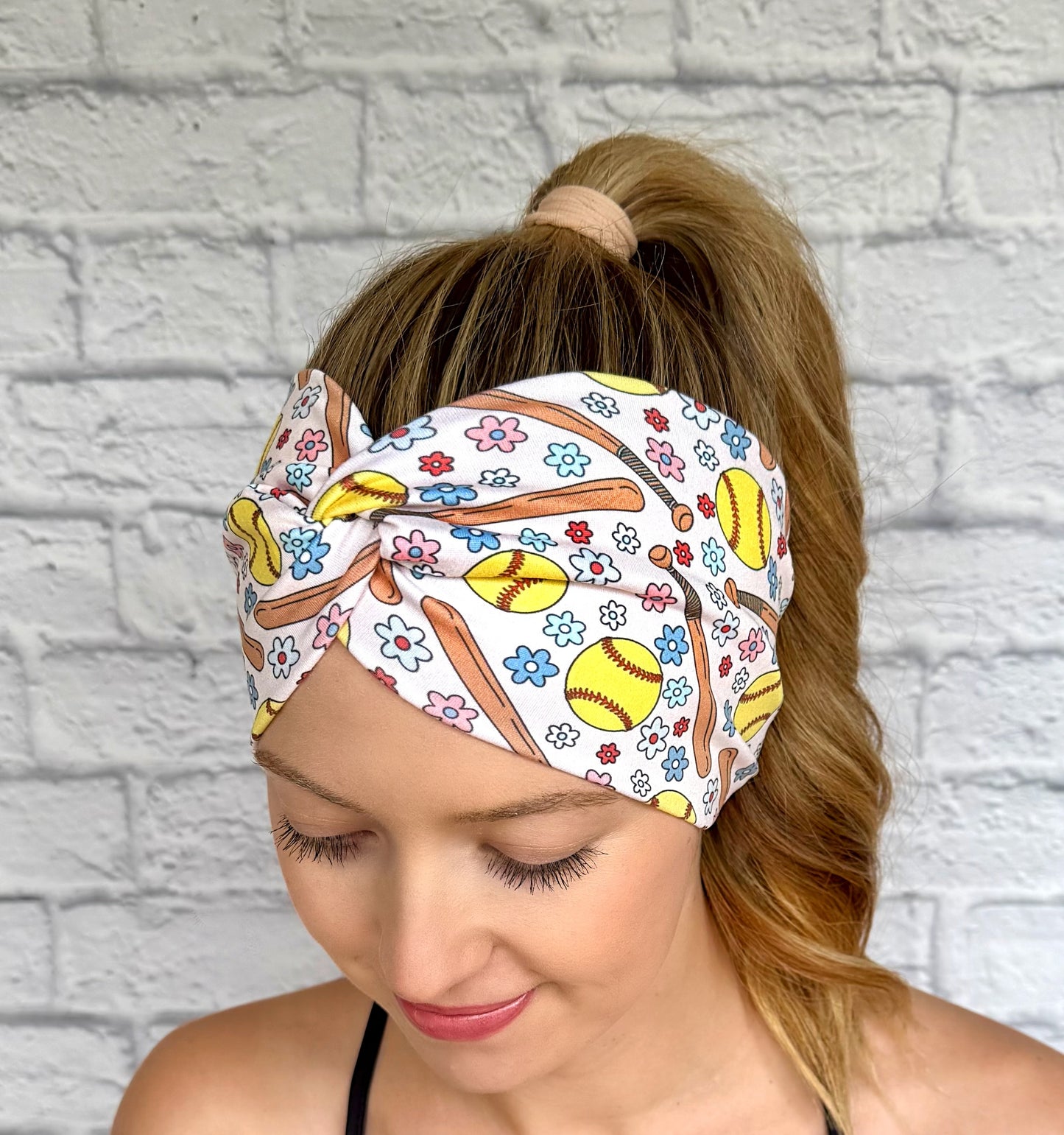 Woman with hair in ponytail wearing wide, twisted headband in cream color with softball and flower theme print.