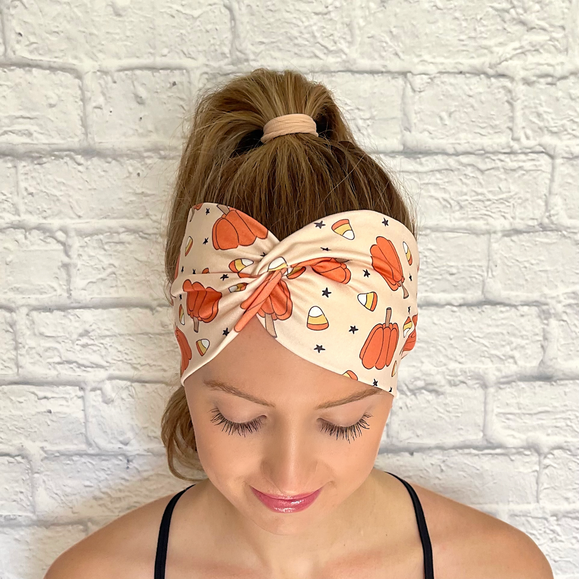 Woman with hair in curled ponytail wearing wide, twisted headband in tan with pumpkin, candy corn, and star print.