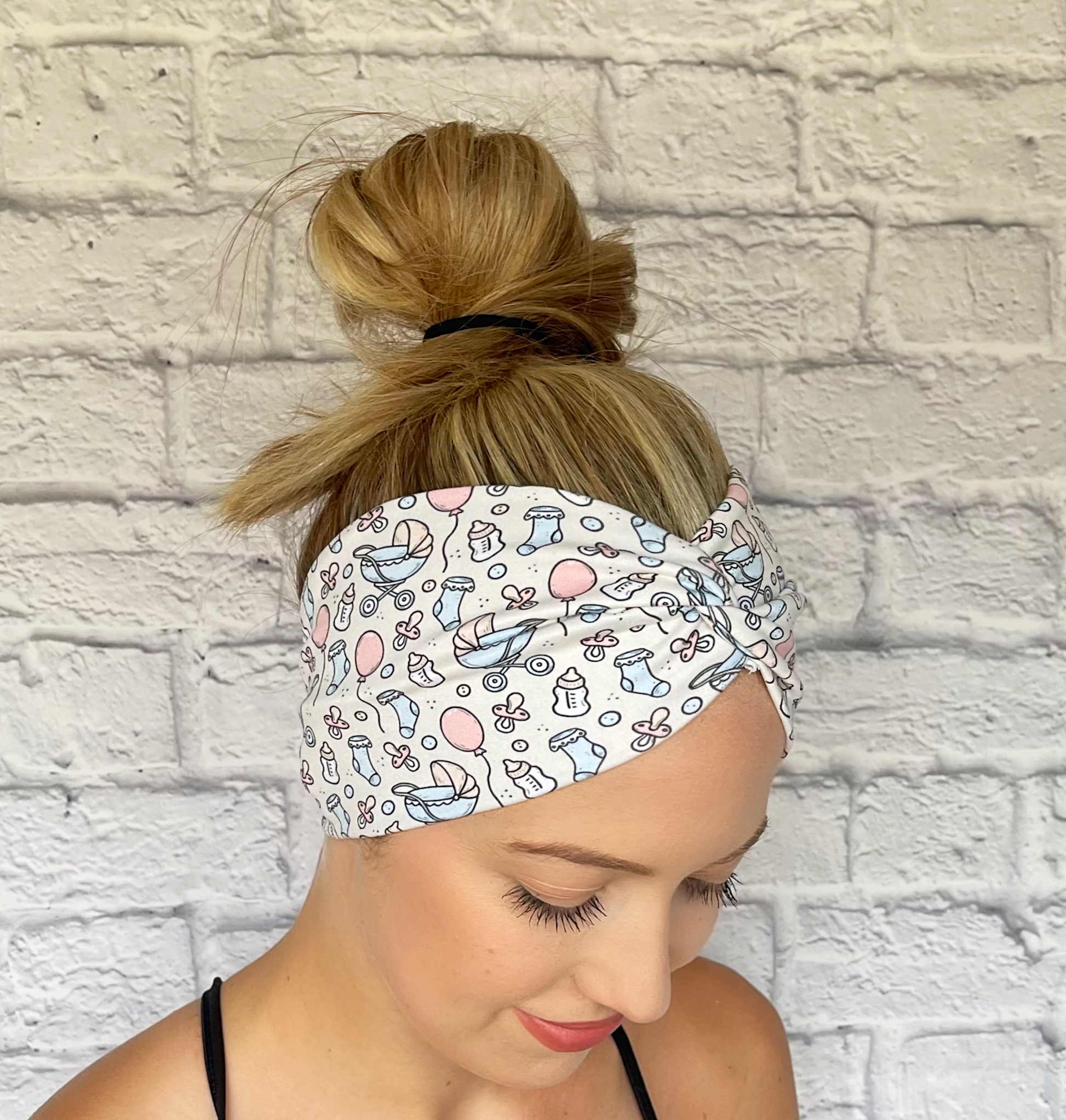 Woman with hair in bun wearing wide, twisted, cream headband with baby theme print- binkies, buggy, socks, bottles, and balloons.