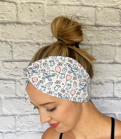 Woman with hair in bun wearing wide, twisted, cream headband with baby theme print- binkies, buggy, socks, bottles, and balloons.