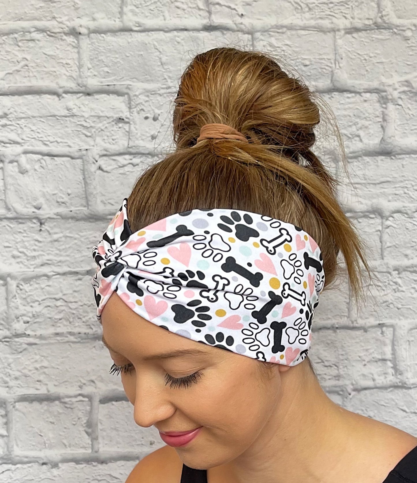 Woman with hair in bun wearing wide, twisted headband in white with black, pink, yellow, and light blue dog theme print with paws, dog bones, and hearts.