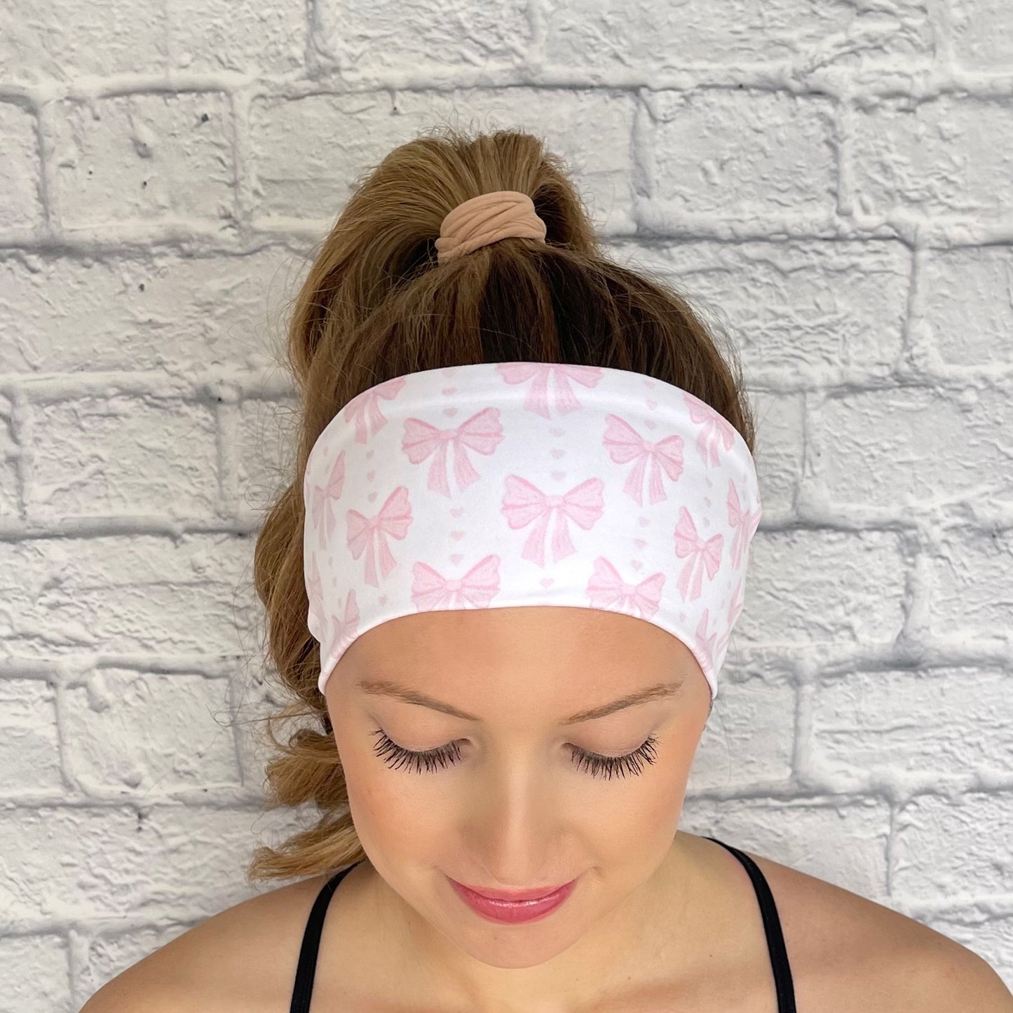 Woman with hair in curled ponytail wearing flat, stretchy, white headband with light pink bow print.