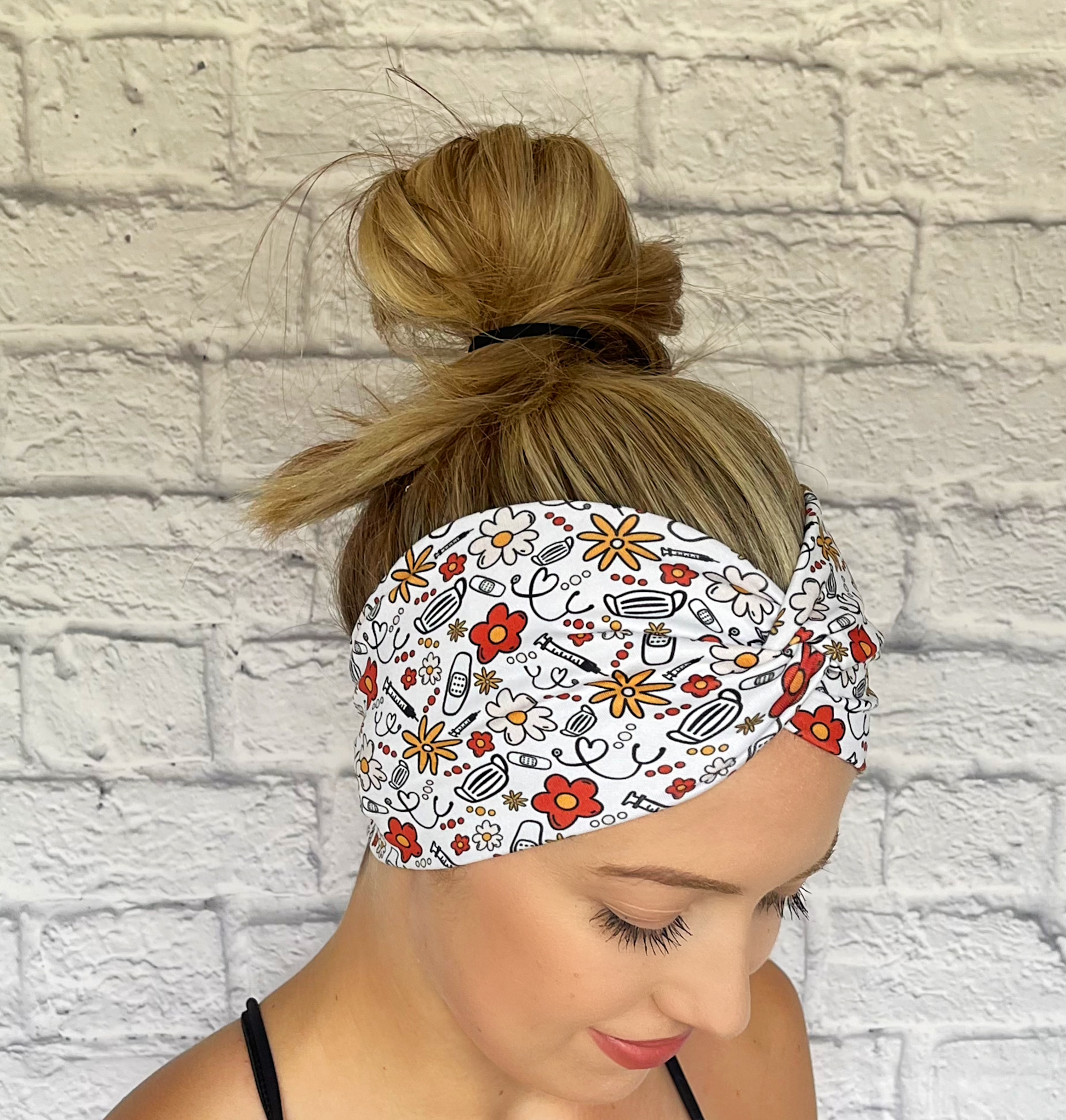 Woman with hair in bun wearing wide, twisted headband in white with red, yellow, and black floral and medical theme print.