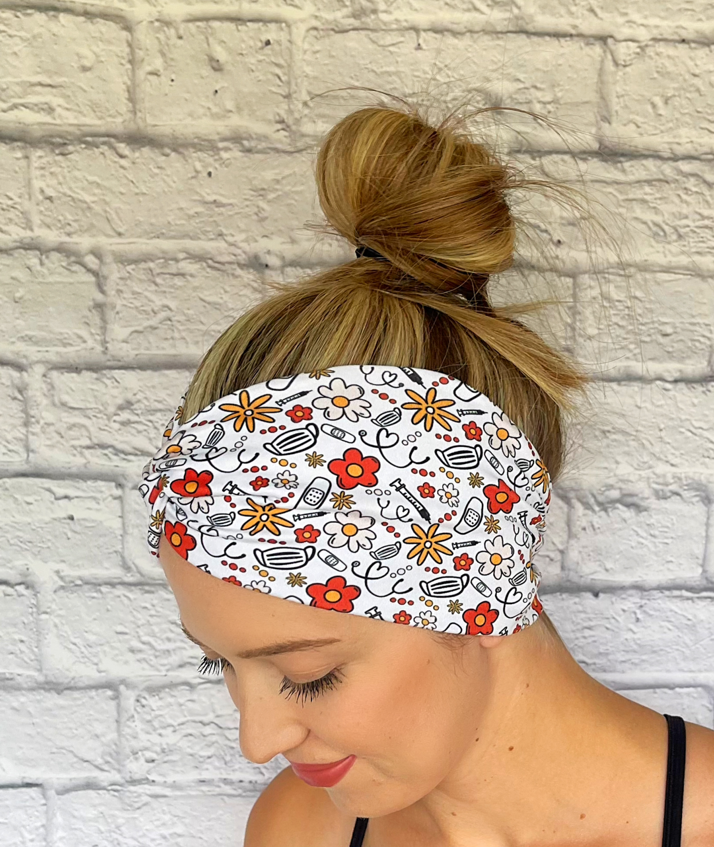 Woman with hair in bun wearing wide, twisted headband in white with red, yellow, and black floral and medical theme print.