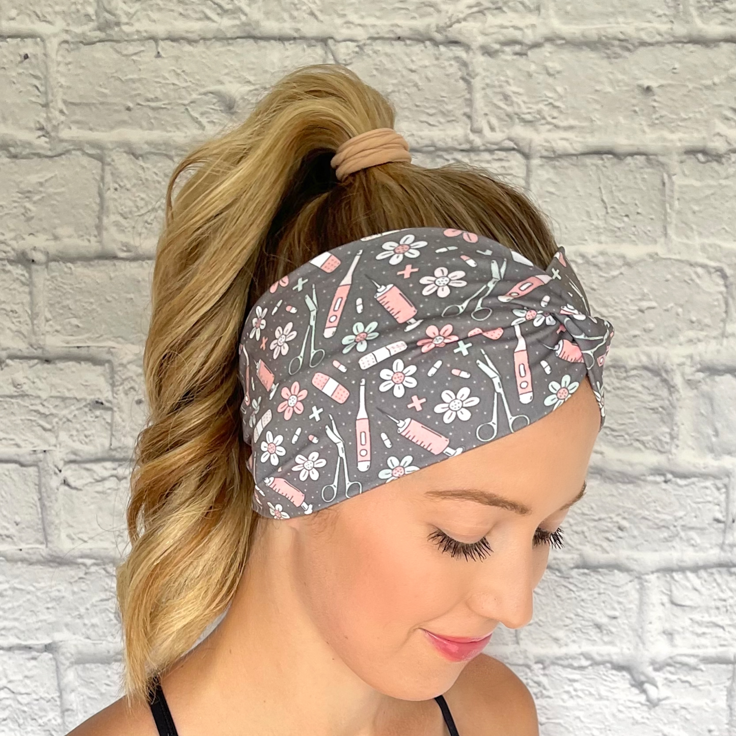 Woman with hair in curled ponytail wearing gray, wide, twisted headband with pink, mint blue, and white medical print.